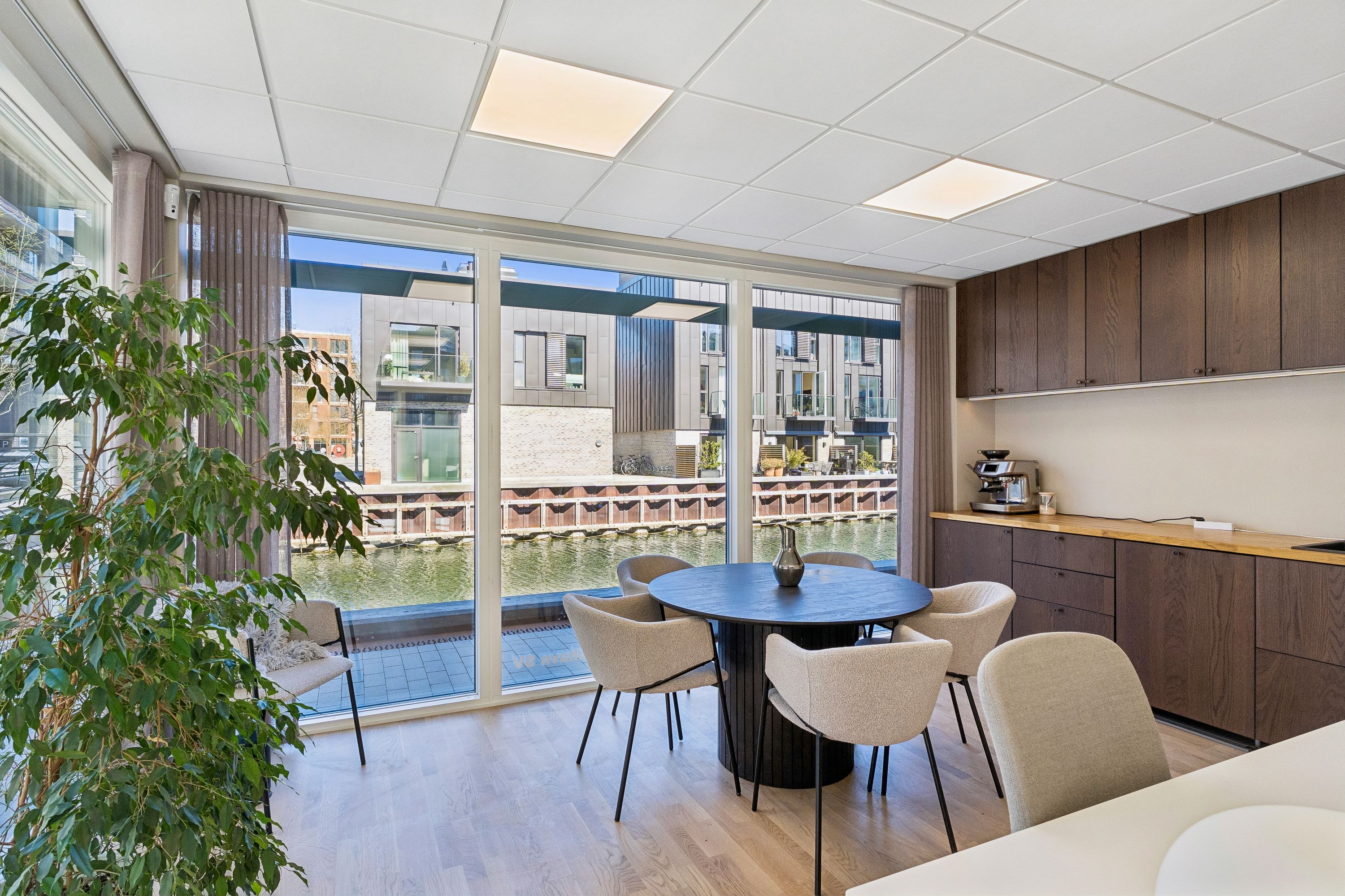 Indkøbsområde med møbelgruppe og plante, højt loft og store ruder med udsigt over vandløb. Køkken med kaffemaskine og lyse træskabe.