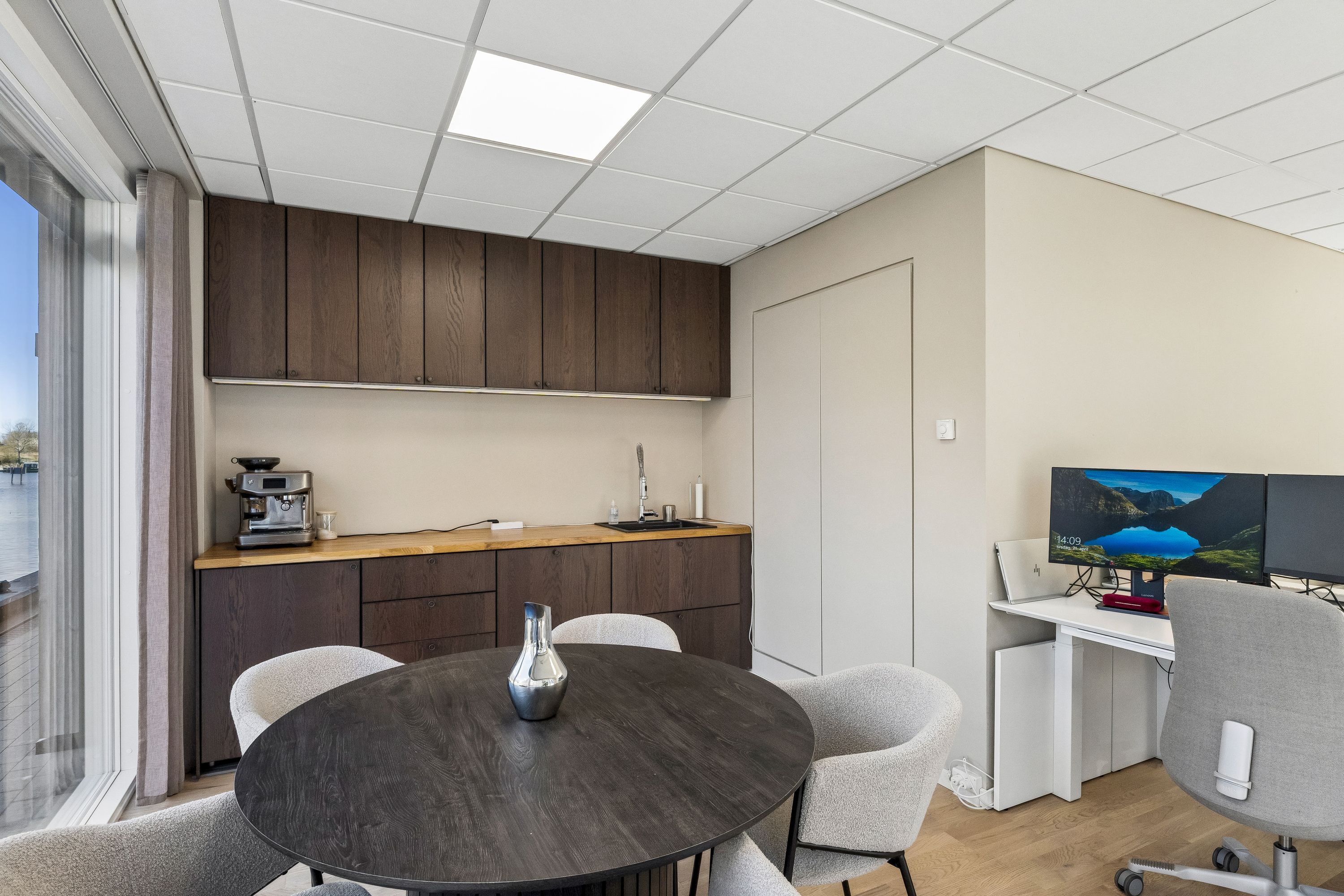 Kontor med skrivebord, computere, skrivebordslamper, skrivebordsvase og kaffemaskine på en trægulv med en rundt bord og sæder