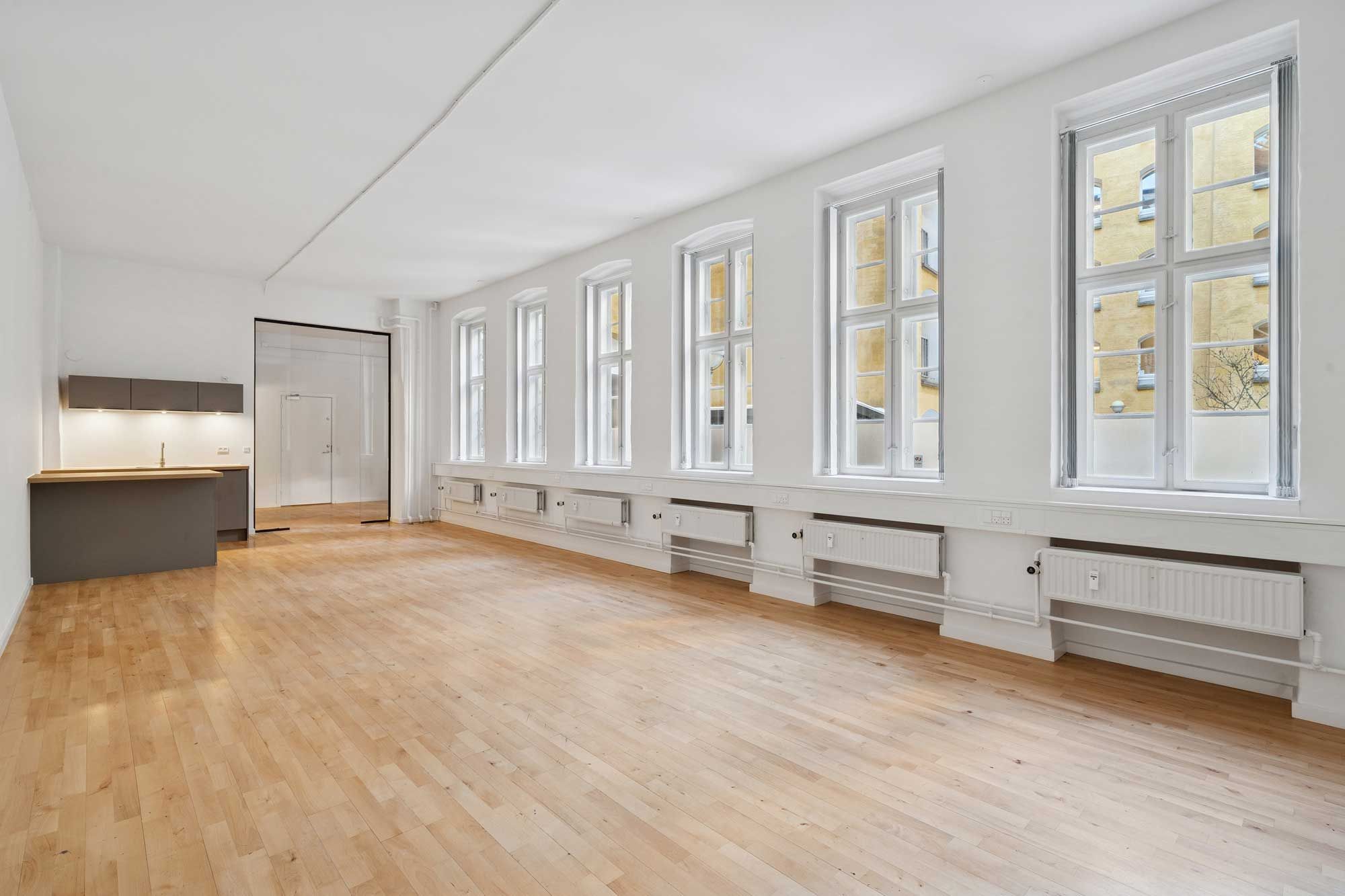 Hvidt kontorlokal med store vinduer og moderne radiatorer, blanke trægulve og et køkken med lyse lofter.