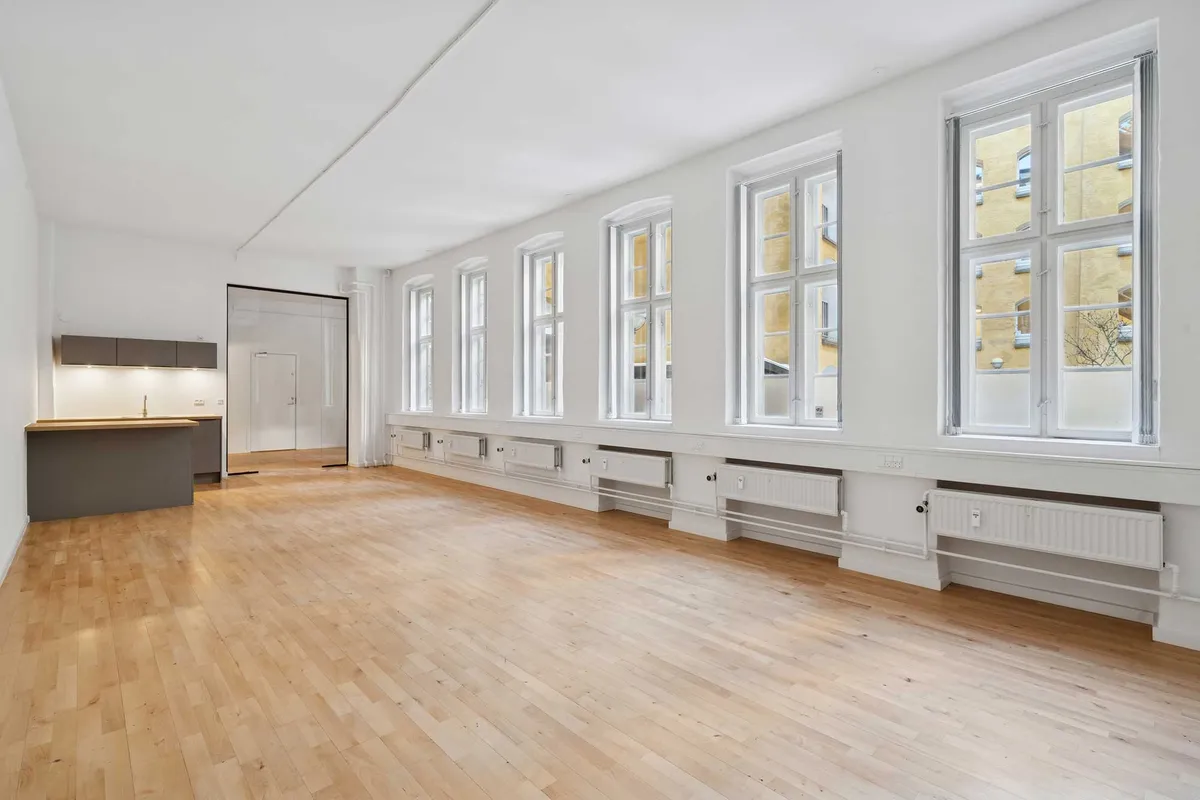 Hvidt kontorlokal med store vinduer og moderne radiatorer, blanke trægulve og et køkken med lyse lofter.