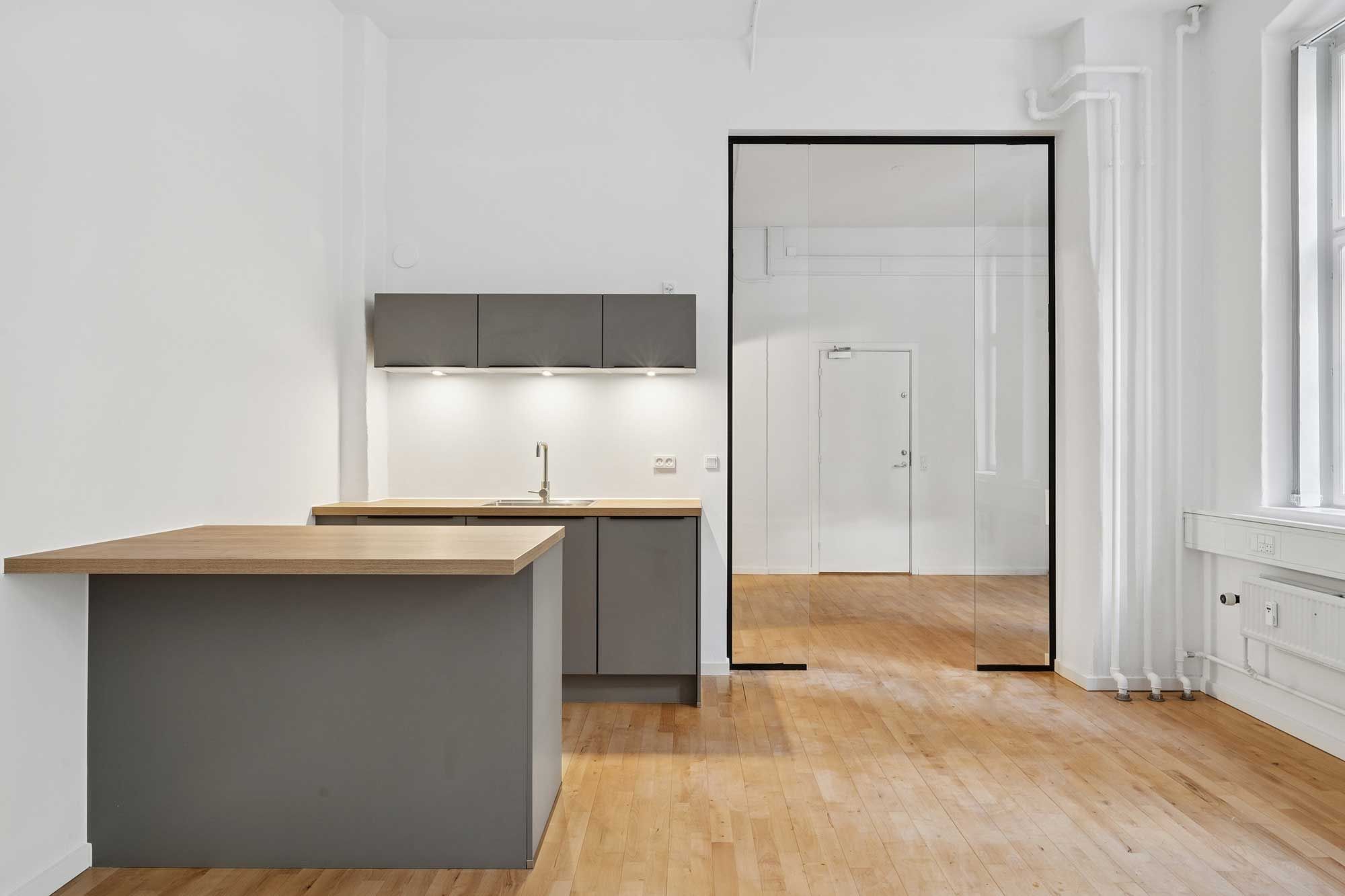 En moderne og minimalistisk køkken med en hvid væg og en trægulv. Der er et søjledesign med et spejl og en dør i baggrunden. Der er en træbar og en grå skabskabinet med en vandhane og et vaskebækken.