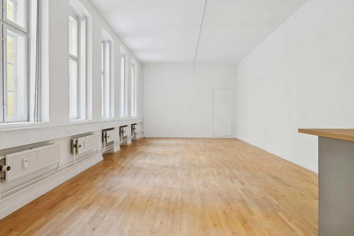 Rummelig stueplan med lyse trægulve og hvide vægge. Vinduer med lysegardiner og radiatorer langs væggen. Et træbord til højre og et hvidt dørkarkas i midten.