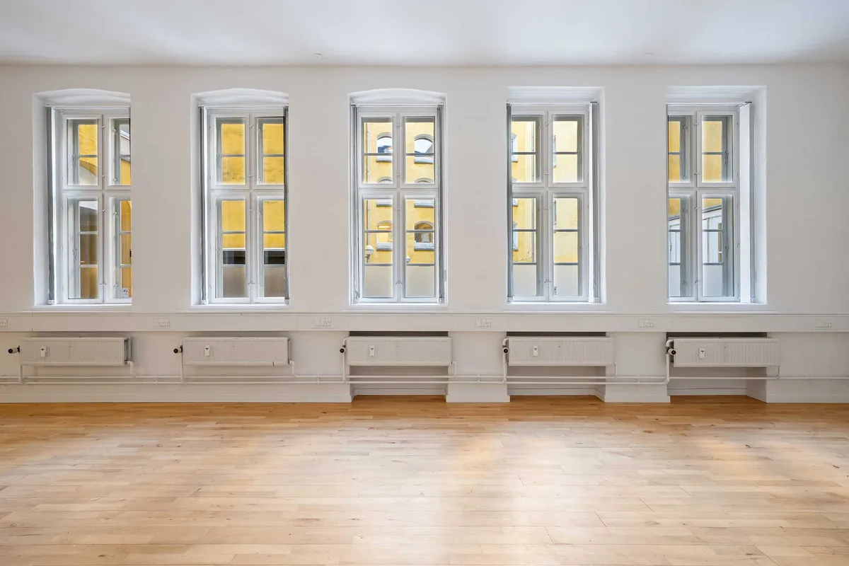 Et tomt kontorlokal med hvide vægge, gulve og fire vinduer med hvide rammer og gule gardiner. Der er fire radiatorer under vinduerne.