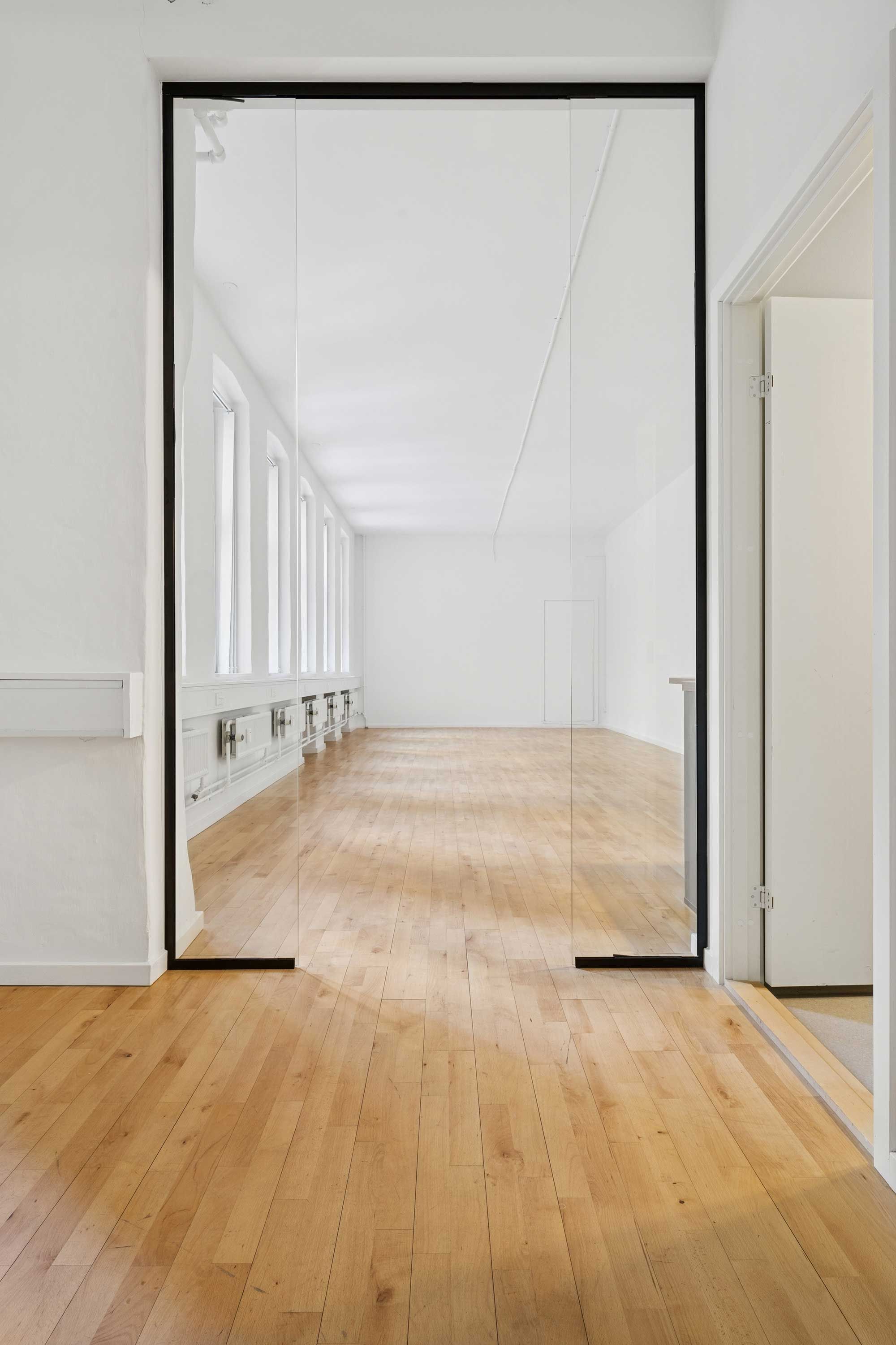 Glasvæg og højloftet rum med lyse trægulve og hvide vægge. Detaljer med gulve og møbler og lyskarv og spejlrefleks.