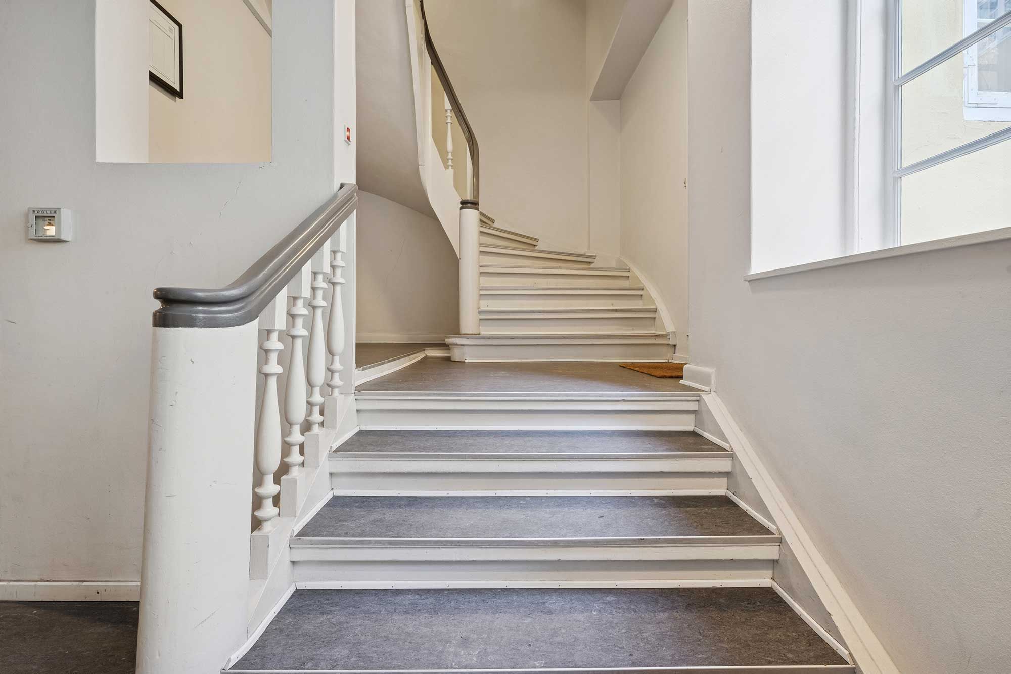 Elegante trapper i et historisk bygning. Væggene er hvide, og der er et vindue til højre. Der er et spejl, der reflekterer et billede. Der er en gulvbelægning og en håndtag på trappen