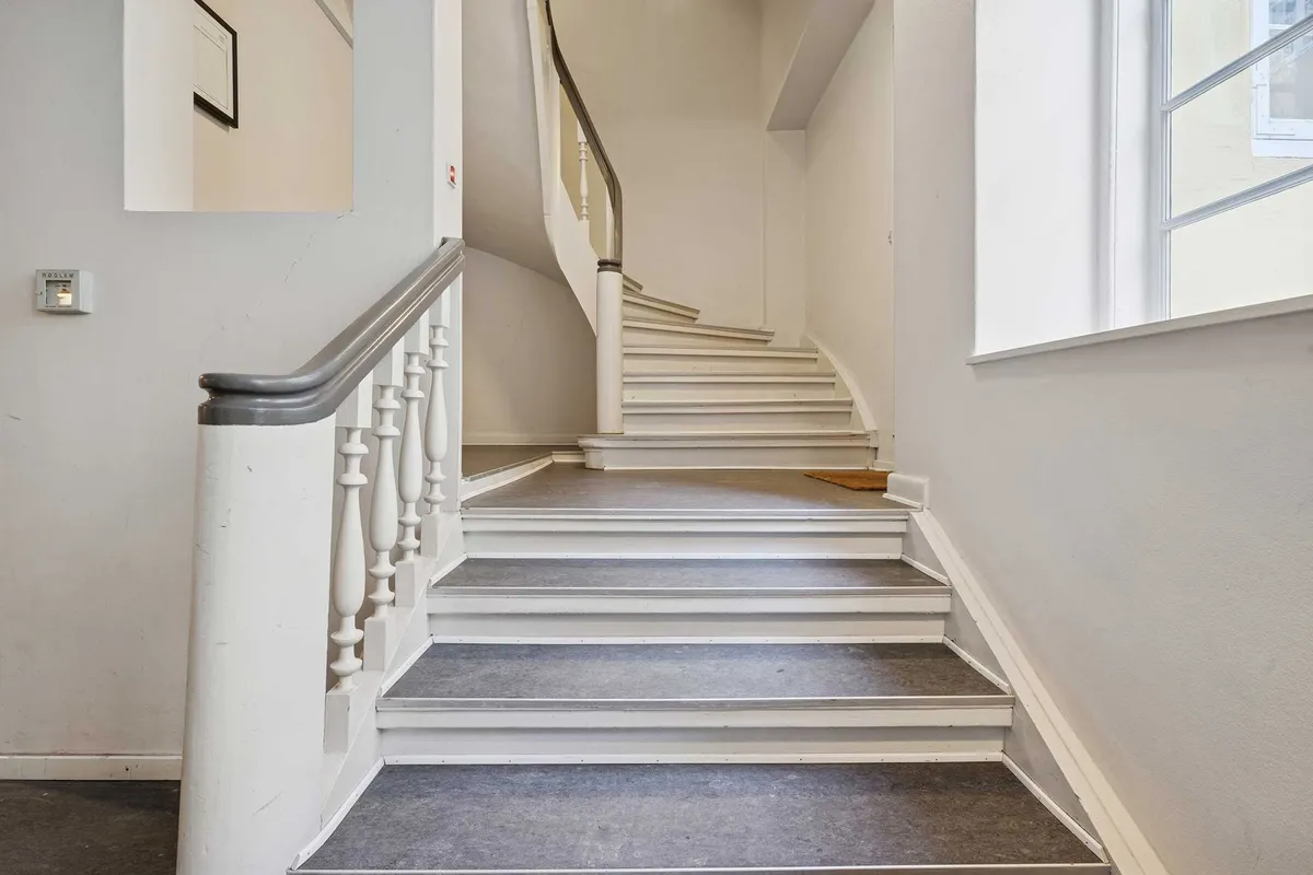 Elegante trapper i et historisk bygning. Væggene er hvide, og der er et vindue til højre. Der er et spejl, der reflekterer et billede. Der er en gulvbelægning og en håndtag på trappen