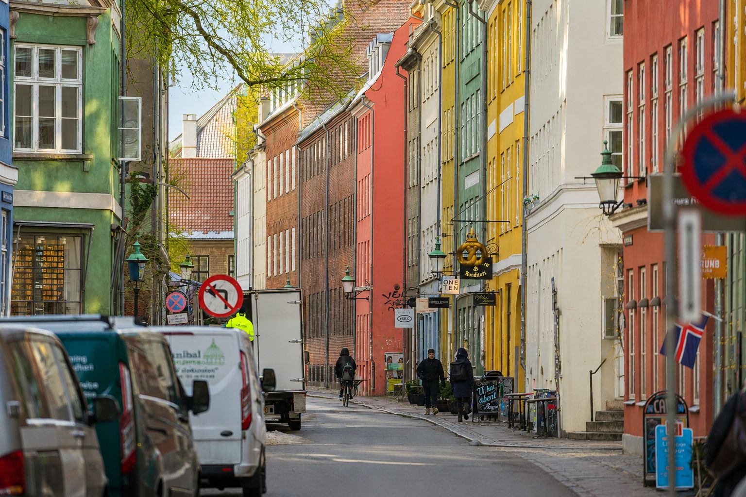 Strædet med farverige bygninger, hvor to personer går på fortovet, og en cykelkører passerer en hvid lastbil og flere parkerede biler.