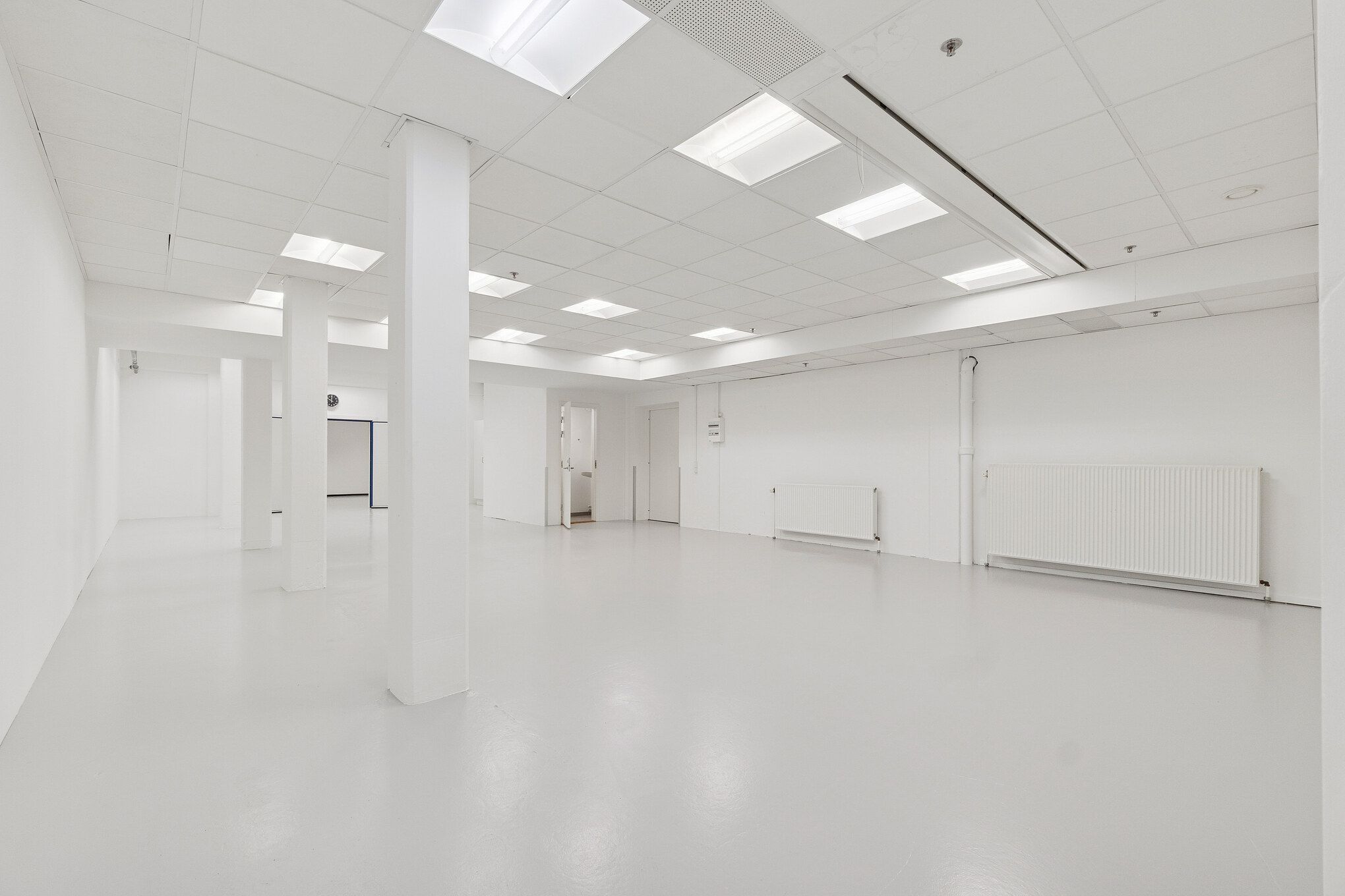 Lager- og showroomlejemål med hvide vægge, blanke gulve, hvide lofter med indbygget belysning og radiatorer. Der er døre og vinduer i rummet.