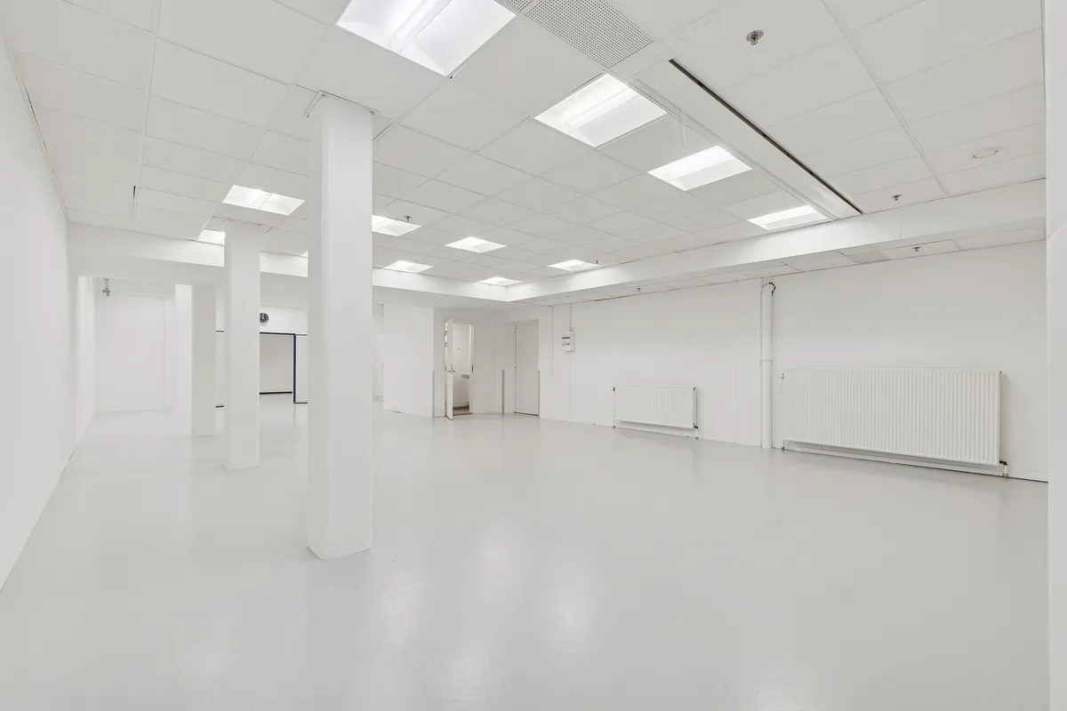 Lager- og showroomlejemål med hvide vægge, blanke gulve, hvide lofter med indbygget belysning og radiatorer. Der er døre og vinduer i rummet.