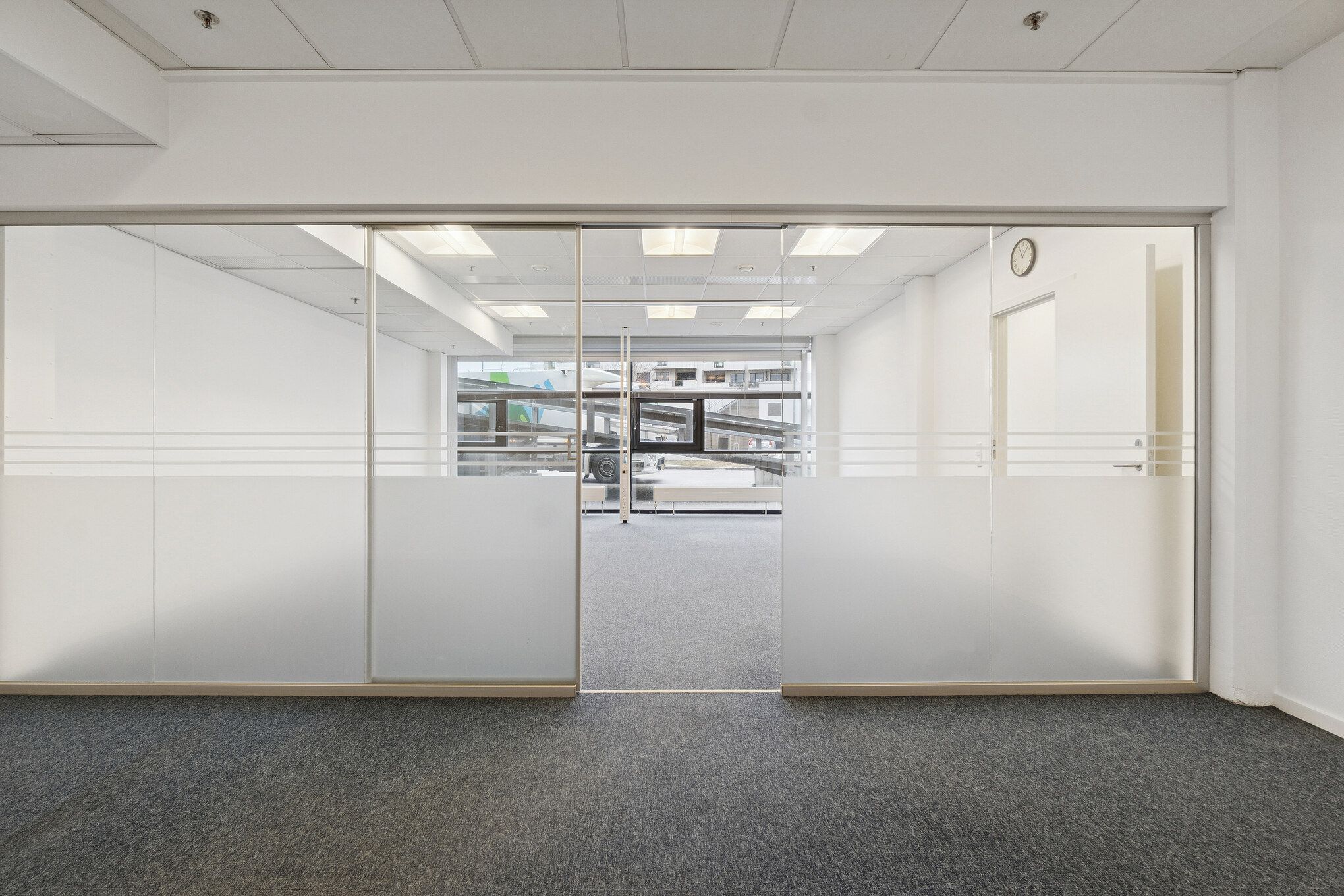 To store glasdøre med metalliske rammer i et moderne kontorrum. Væggene er hvide og gulvet er dækket med gråt gulvtæppe. Loftet er hvidt og indeholder flere lysekure.