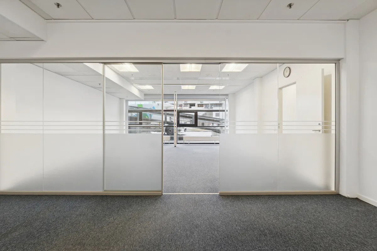 To store glasdøre med metalliske rammer i et moderne kontorrum. Væggene er hvide og gulvet er dækket med gråt gulvtæppe. Loftet er hvidt og indeholder flere lysekure.