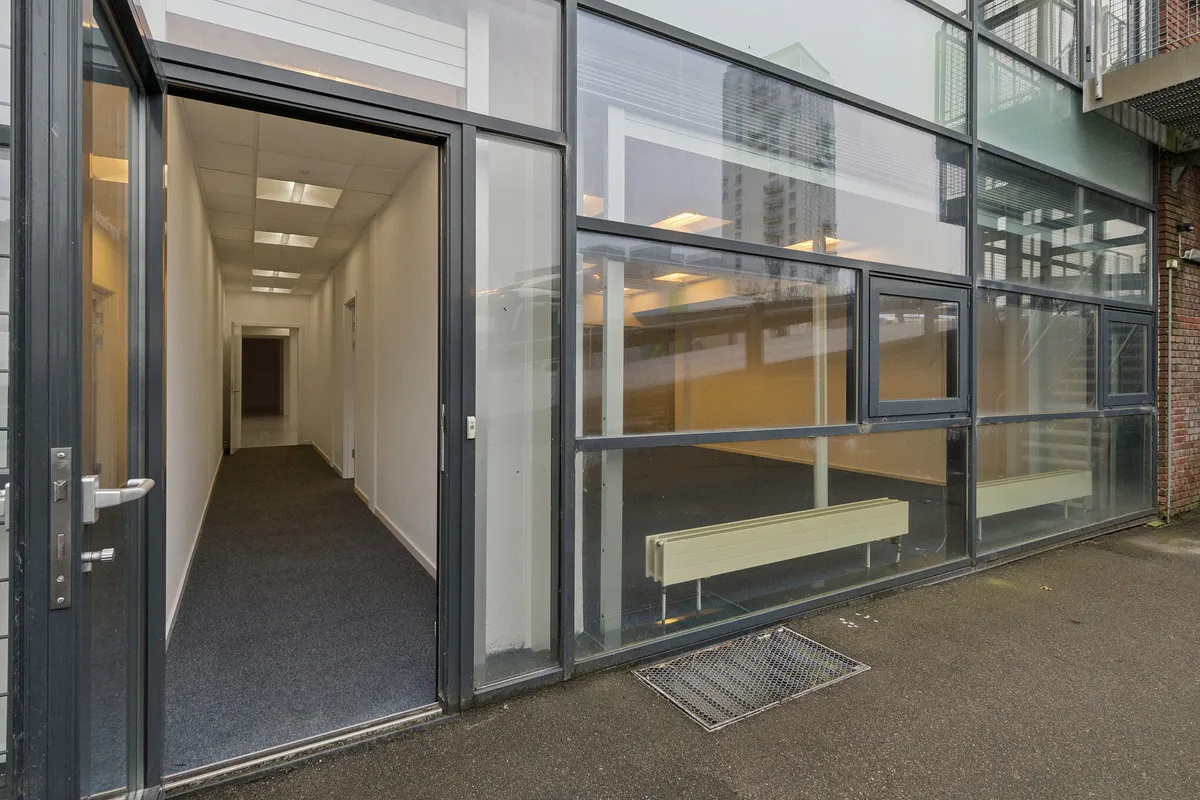 Udenfor en moderne bygning med store glasruder er der en indgang med dobbeltdøre. Lejligheden har en åben gang med gulvbelægning og refleksioner af bygningen. Der er radiatorer på gulvet og gulvvarme samt et skraldkar.