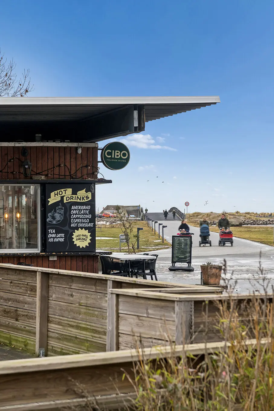 Café Cibo med solrige veje og stolpebord, hvor folk kører med vogn og gåstier i baggrunden under en klar himmel