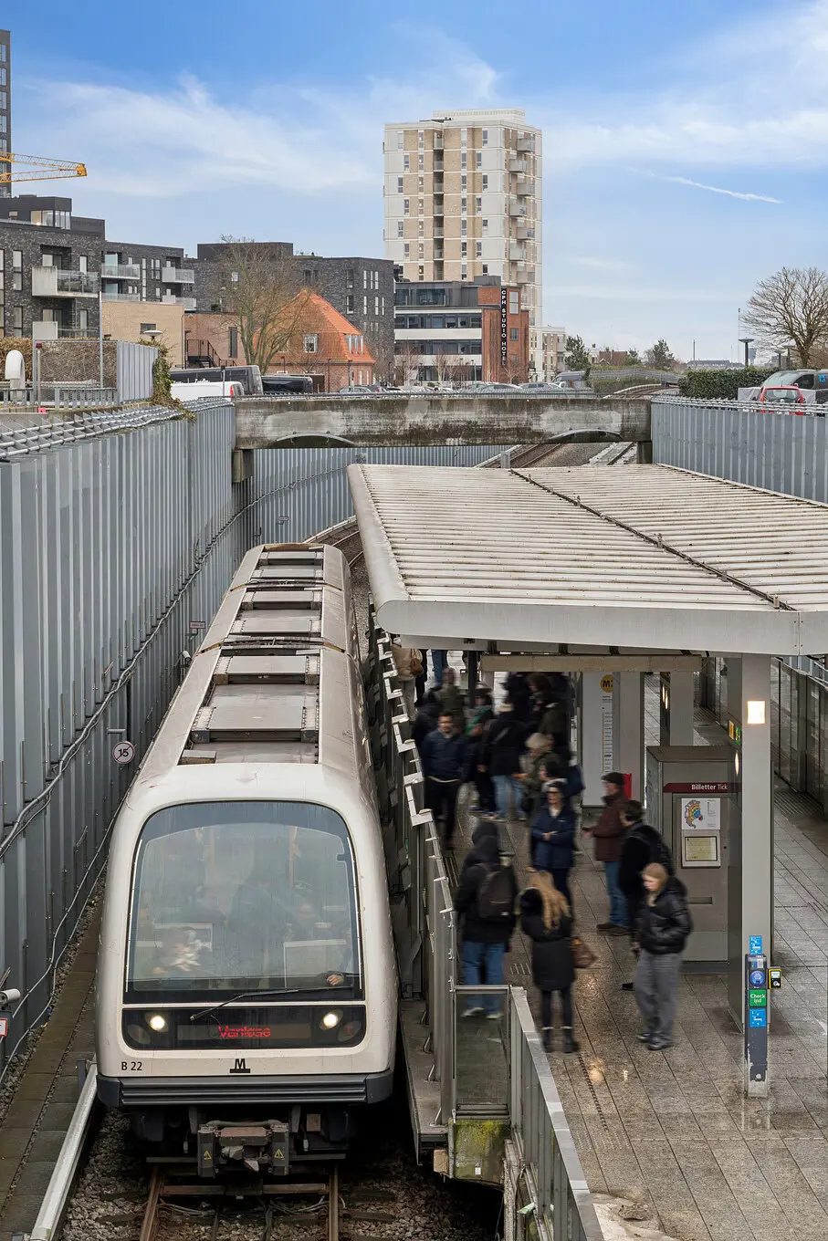 Metrostation med hvid metrovogn og flere passagerer. Stationen er dækket af et tag og ligger ved siden af en jernbanebro. Bygninger og træer i baggrunden.