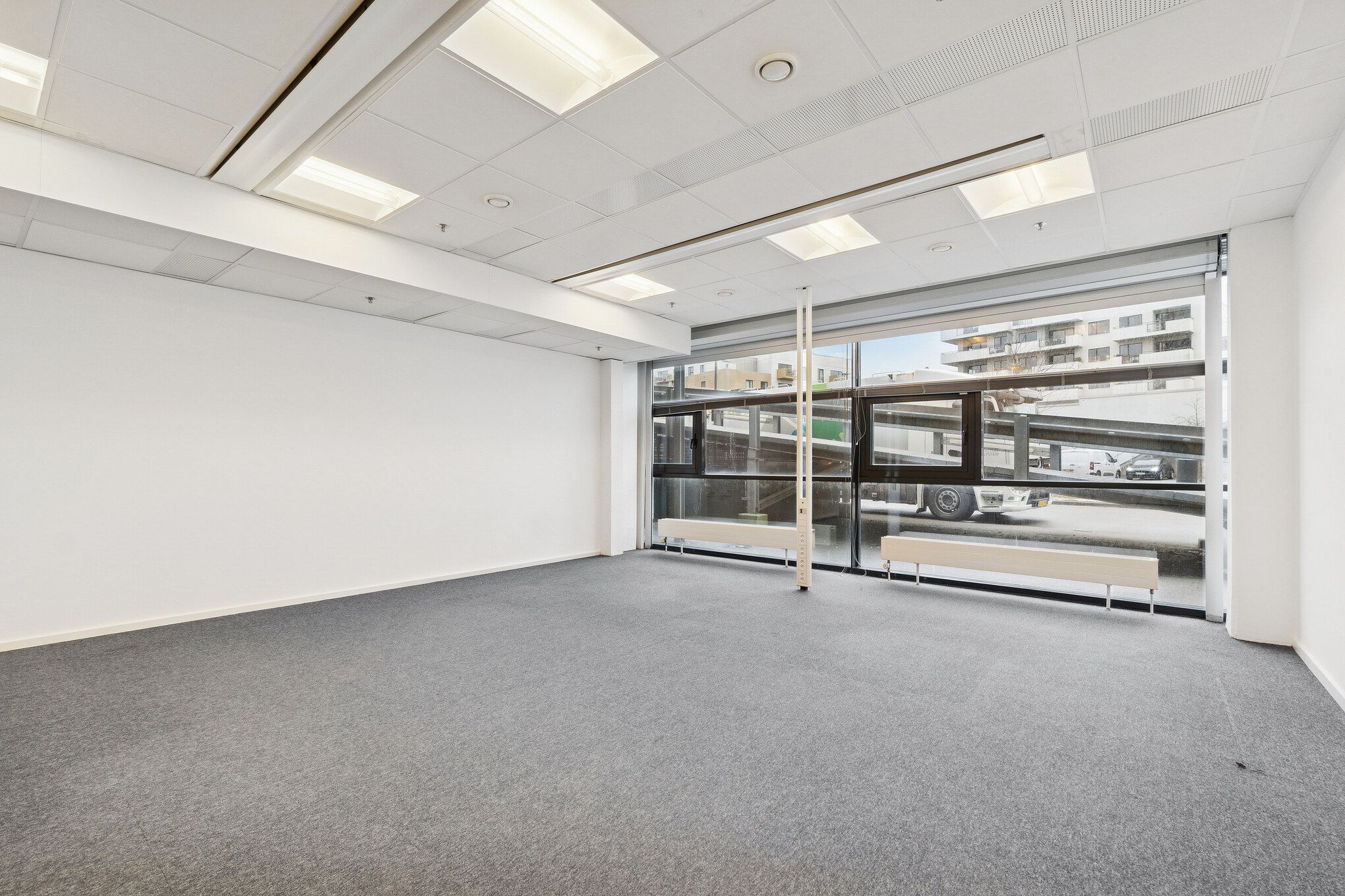 En stor, glasklædt lejlighed med moderne indretning og et vidt udsigtsvindue mod gaden og en parkeringsplads med biler og busser. Rummet er møbleret med to lange, hvide radiatorer og en lysegrå gulvbelægning.