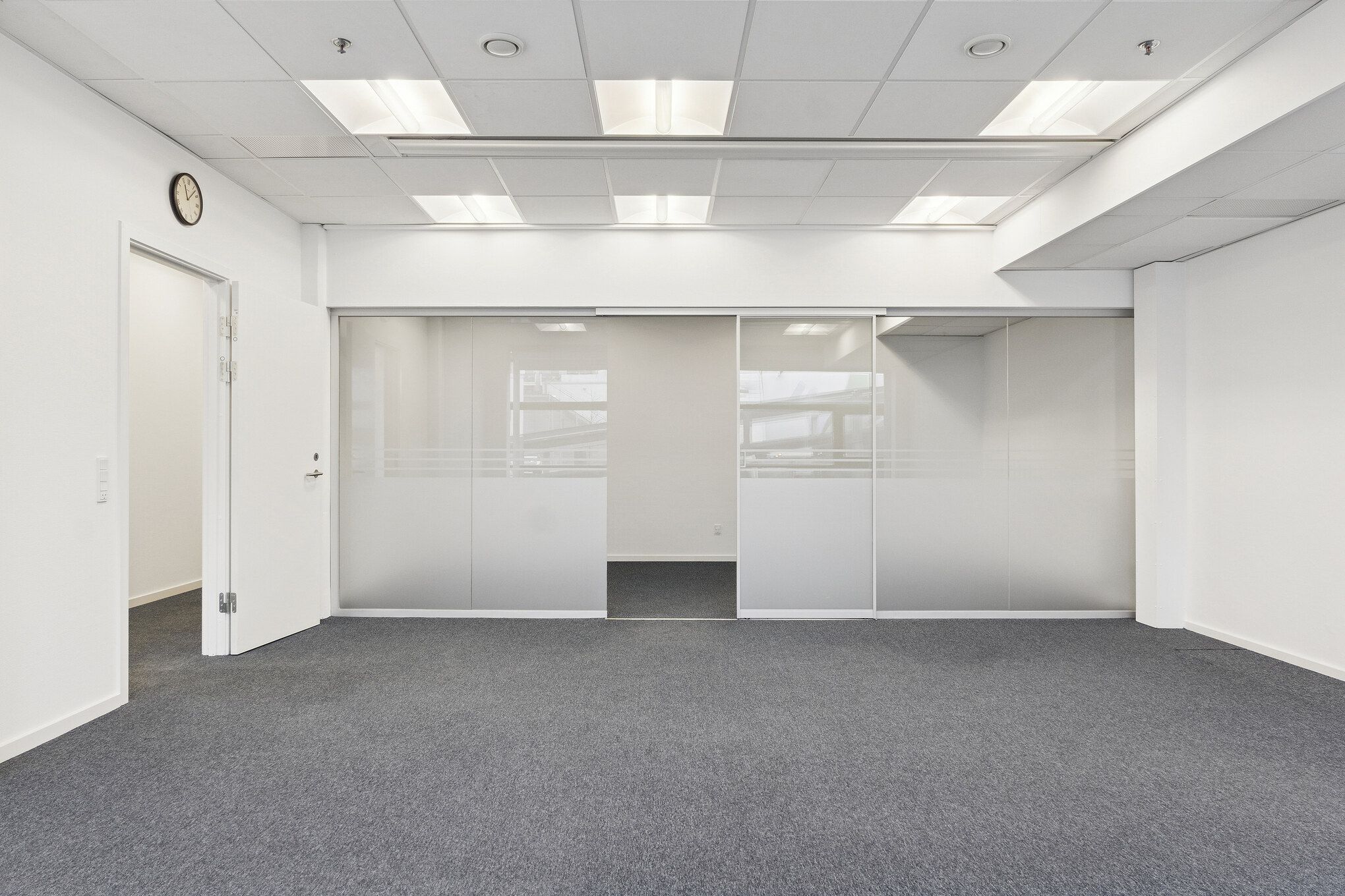 Et stort og lyset lejemål med hvide vægge og glasdøre, der åbner op mod en lysgrå gulvbelægning. Væggen har et ur, og loftet er udstyret med flere lysarme.