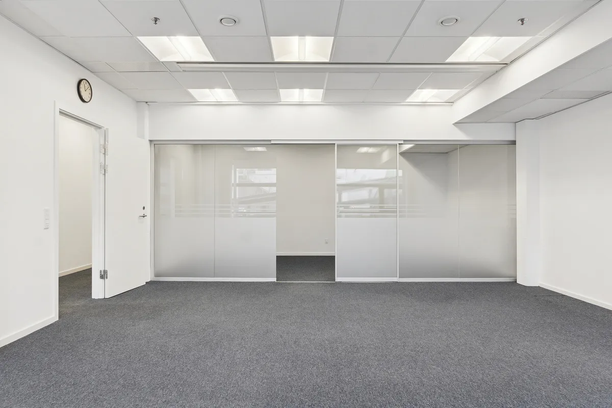 Et stort og lyset lejemål med hvide vægge og glasdøre, der åbner op mod en lysgrå gulvbelægning. Væggen har et ur, og loftet er udstyret med flere lysarme.