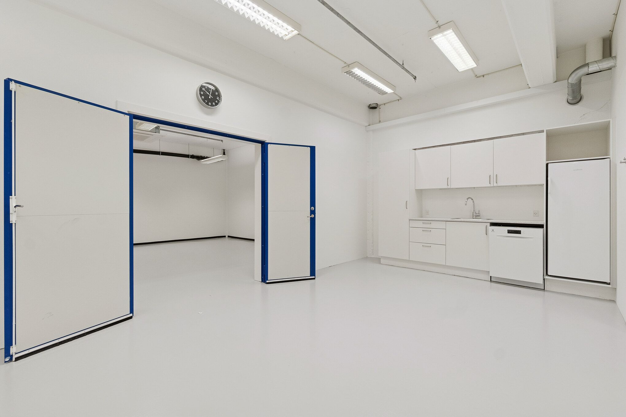 Velindrettet lager og showroomlejemål med hvide vægge, moderne apparater og åbne døre til lagerrum. Indeholder køkken med hvide skabsfronter, lysekroner og et træk til opvaskemaskine.