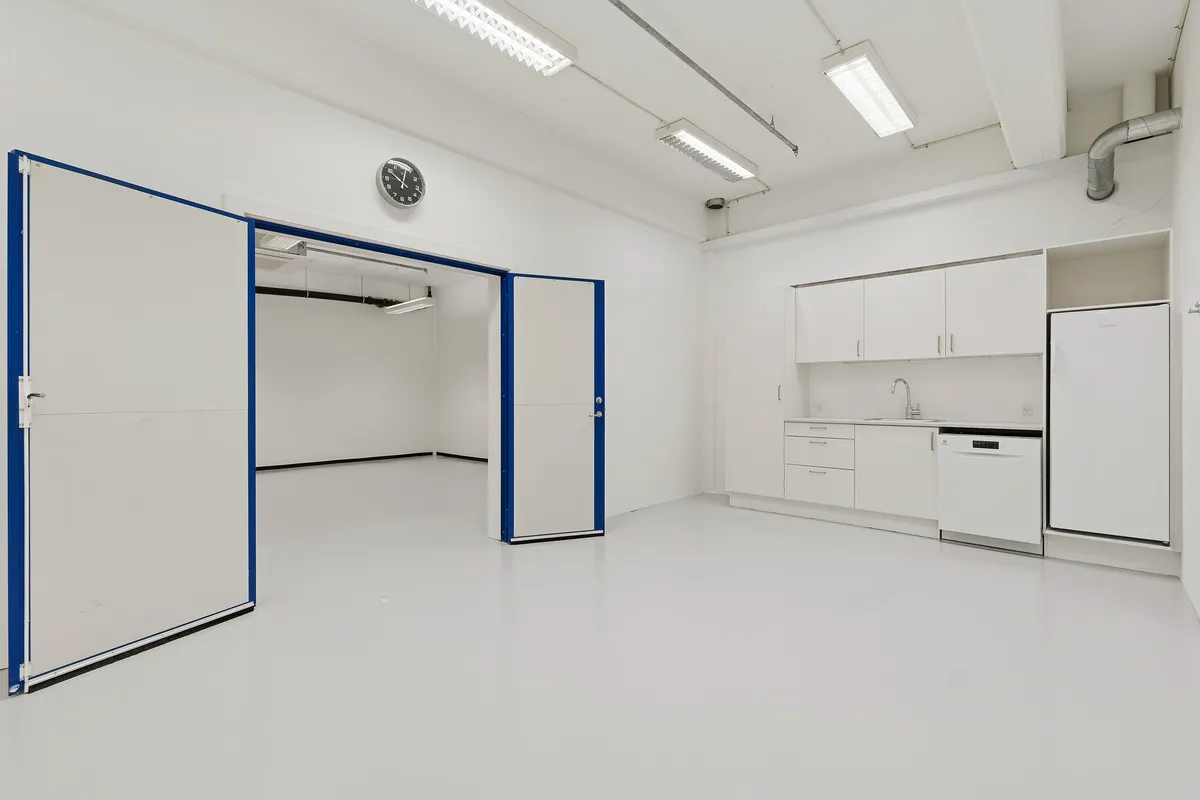 Velindrettet lager og showroomlejemål med hvide vægge, moderne apparater og åbne døre til lagerrum. Indeholder køkken med hvide skabsfronter, lysekroner og et træk til opvaskemaskine.
