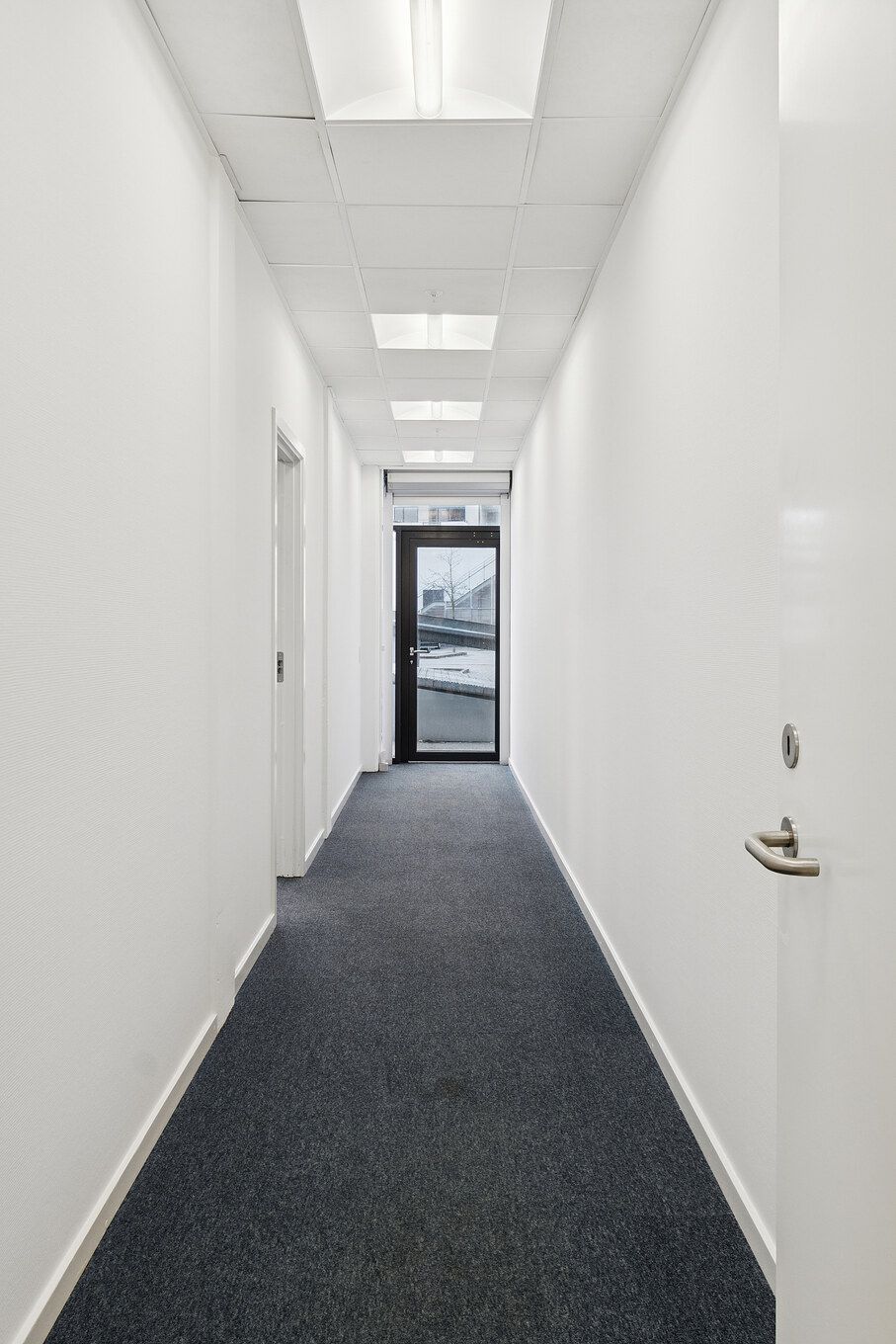 En lang, smal korridor med hvide vægge og mørke gulvbelægning. To hvide døre på højre side og et glasdør med metalramme og sølvfarvet håndtag i enden af korridoren.