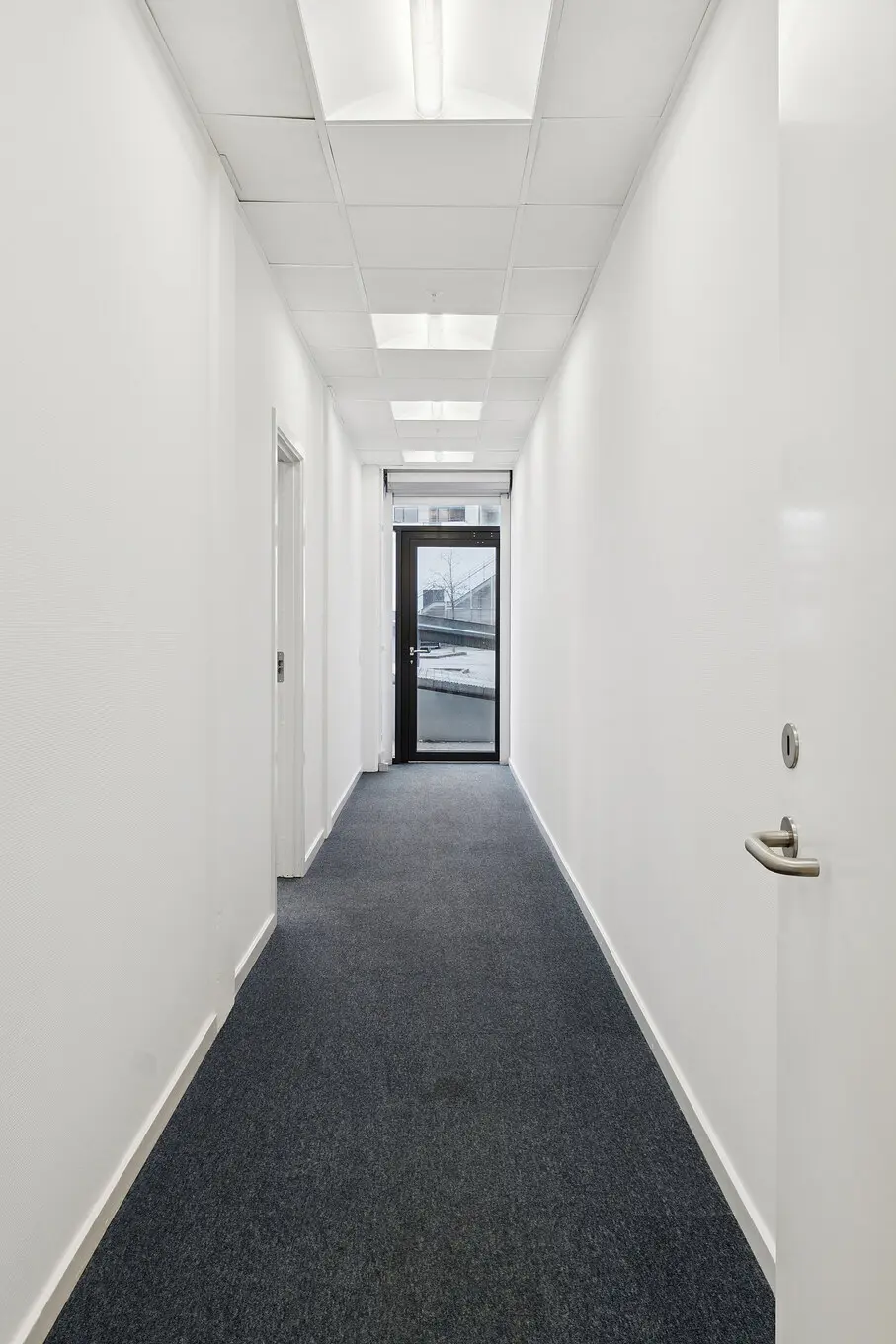En lang, smal korridor med hvide vægge og mørke gulvbelægning. To hvide døre på højre side og et glasdør med metalramme og sølvfarvet håndtag i enden af korridoren.