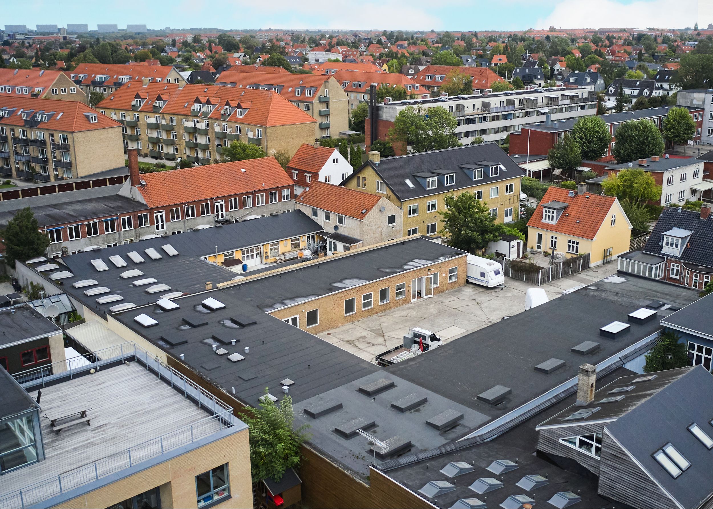 Produktionslokaler med røde tag og moderne arkitektur i Vanløse. Forskudte containere og parkeringsplads. Tæt bebyggelse med røde og gule tag.