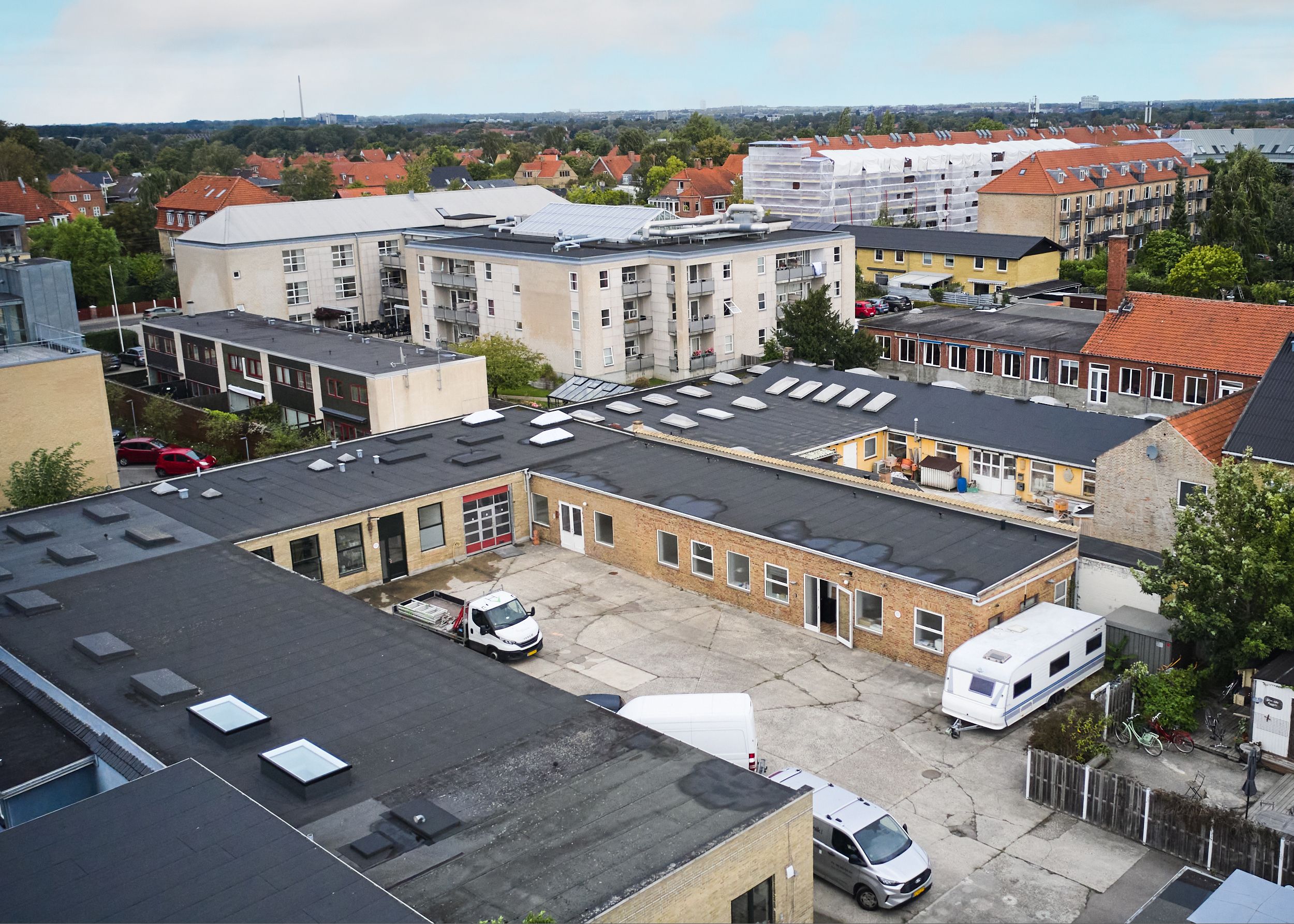Produktionslokaler med flere biler parkeret i parkeringsplads. Huset med røde tag. Solpaneler på tag. Forskellige bygninger i baggrunden. Træer og grønne områder i baggrunden.