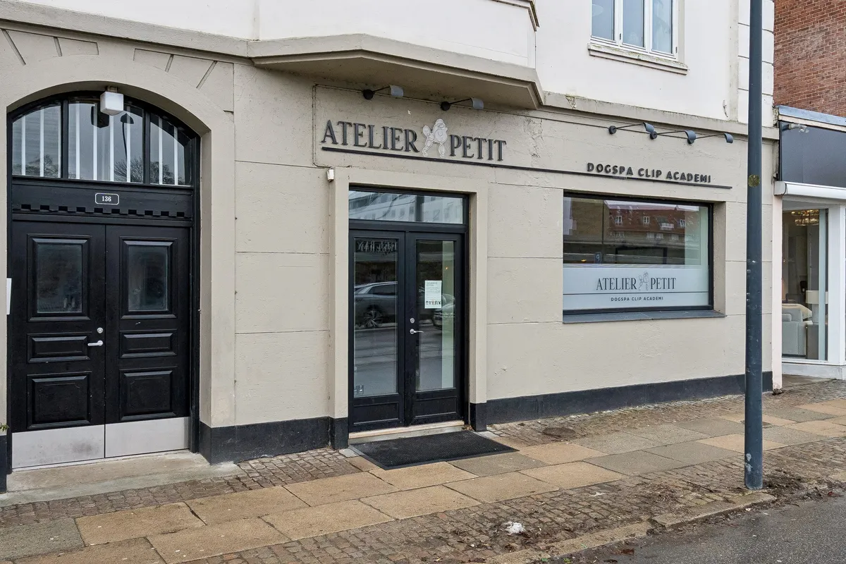 Moderne, hvidt byggeværk med to dobbeltdøre og to butiksruder, begge med skiltet Atelier Petit. Butiksruderne har teksten DogSpa Clip Academi.