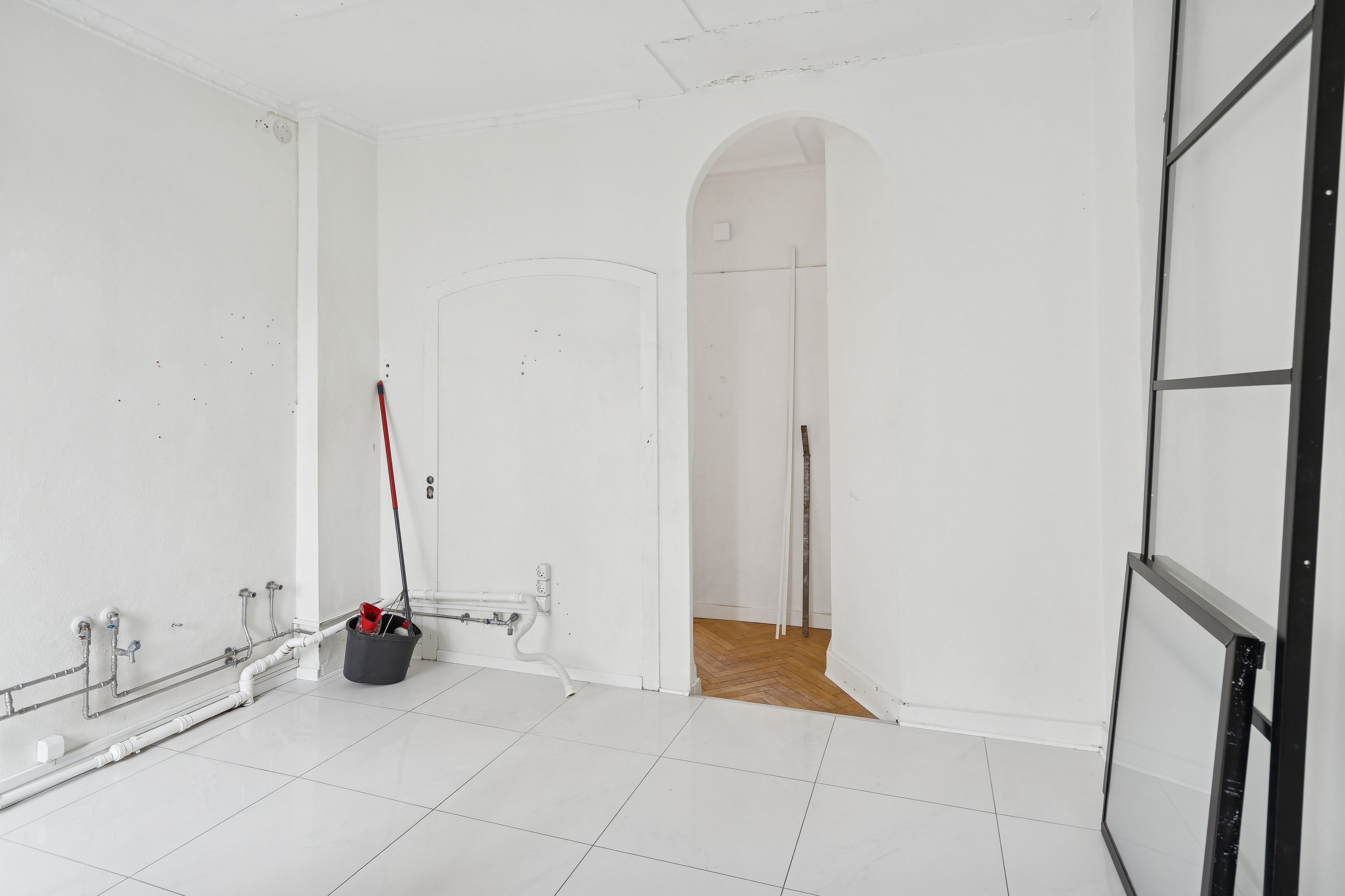 En ren og tom stue med hvide vægge og gulv samt røret på væggen, der er tilsluttet en stålrør. Et skovl og sæk er stillet op i hjørnet.