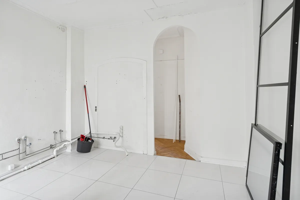 En ren og tom stue med hvide vægge og gulv samt røret på væggen, der er tilsluttet en stålrør. Et skovl og sæk er stillet op i hjørnet.