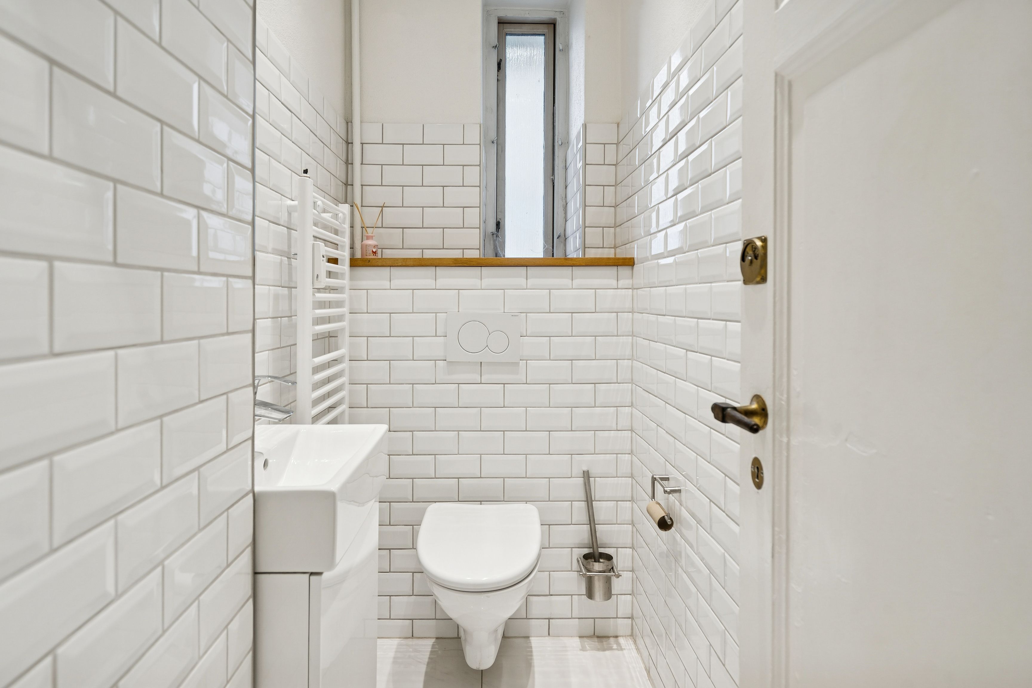 Moderne toiletrum med hvide fliser og lyse møbler. Toilettet med åbne toiletbriller og lukket dør. Hvidt vask og tørretumbler i hjørnet