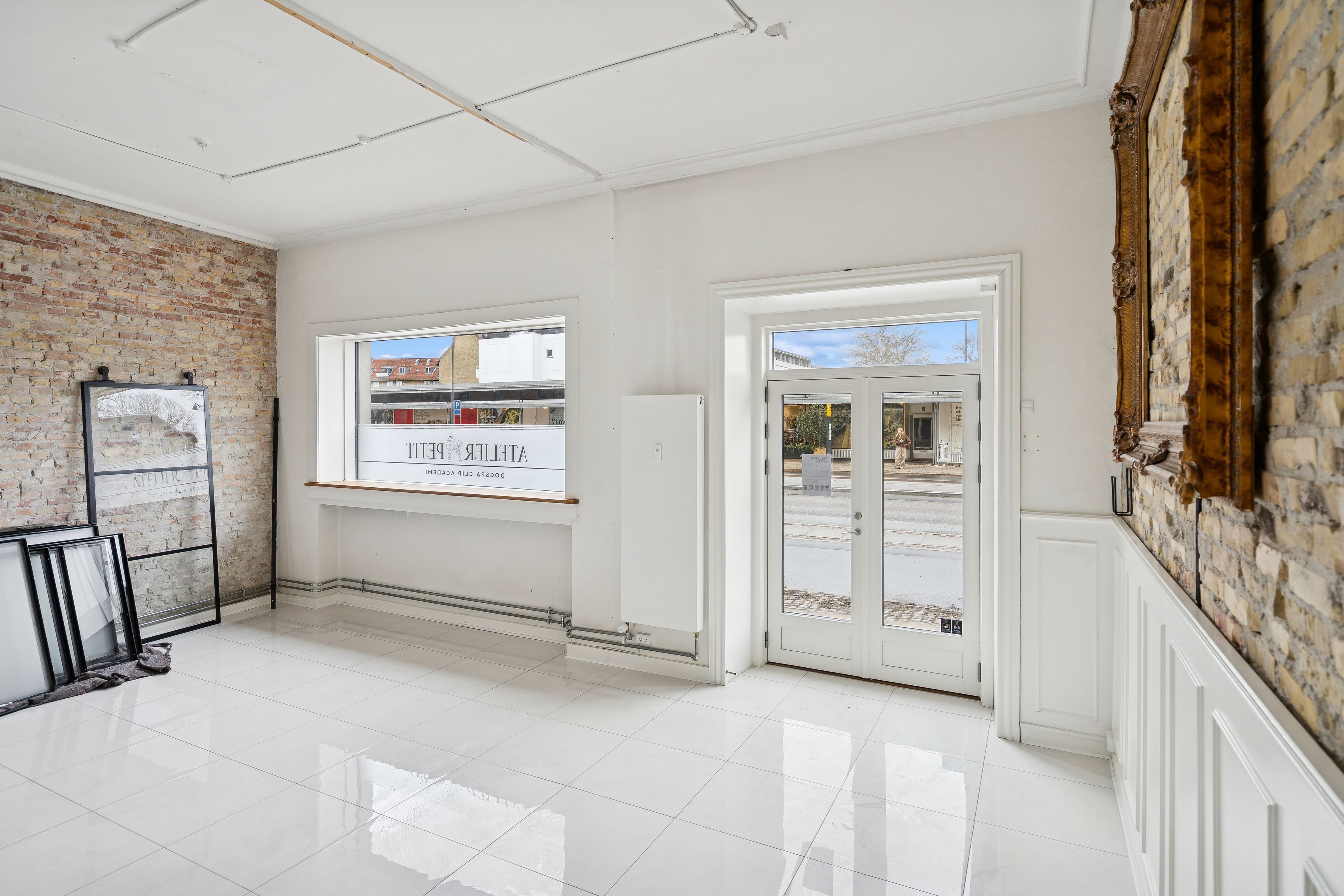 Forretningslokale med hvidt klinkegulv, store vinduer og en dekorativt udsmykket mur. Lyse og moderne interiør med lyse og indbydende rammer.