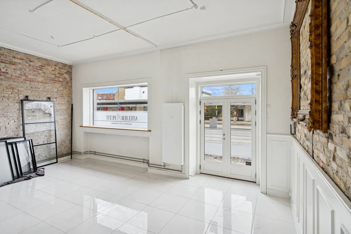 Forretningslokale med hvidt klinkegulv, store vinduer og en dekorativt udsmykket mur. Lyse og moderne interiør med lyse og indbydende rammer.