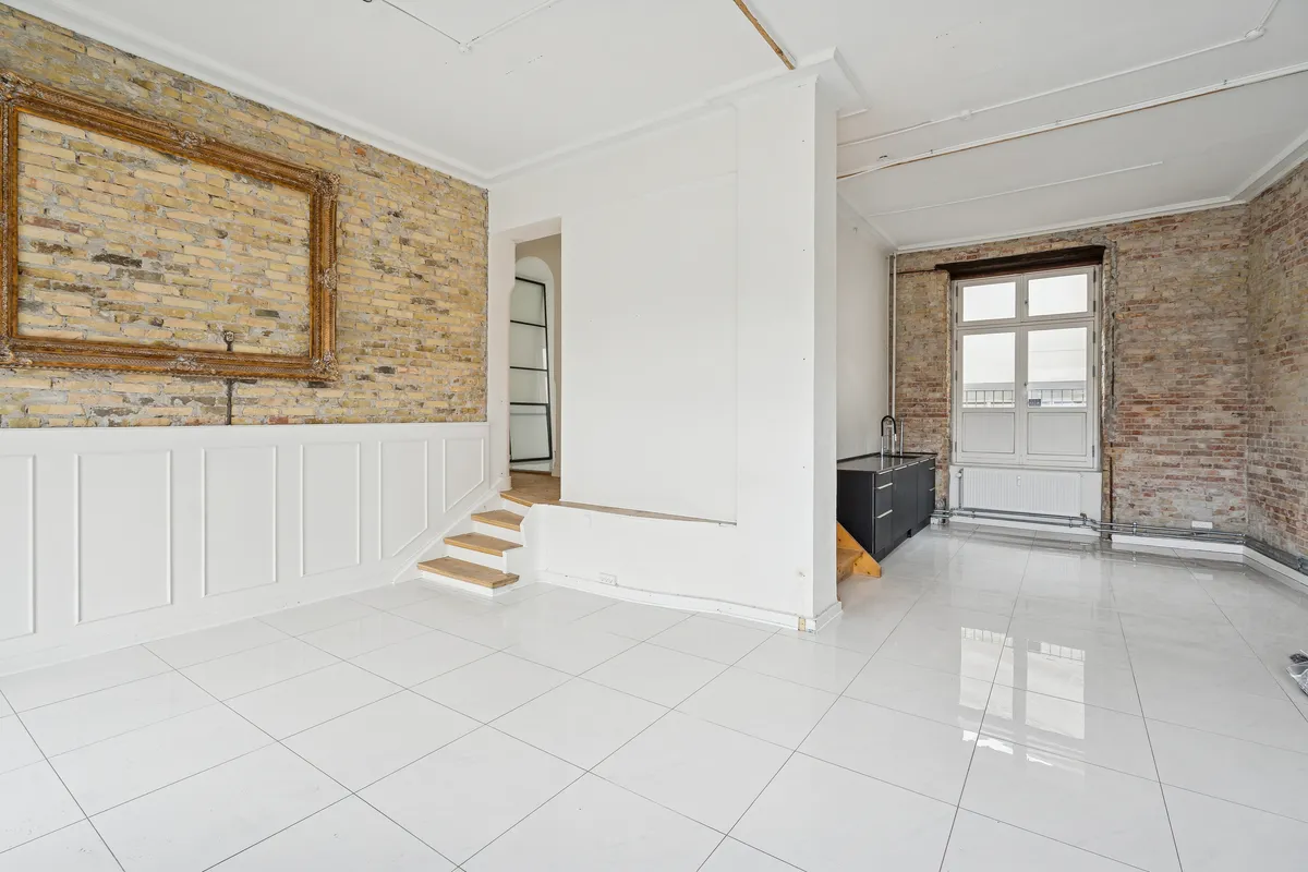 En tom stue med lysegult gulv og to lysegrå vægge med hvide paneler og vinduer. Der er et skab og en stige med trætrin.