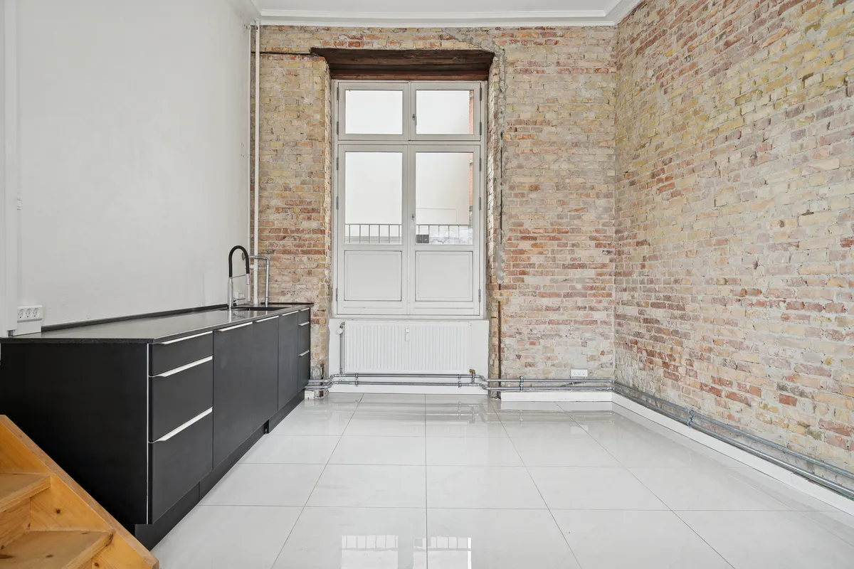 Rum med blanke gulvfliser og et køkken med hvidt og sort design og skraldespand samt radiator og et vindue med hvide ruder og træindfatning