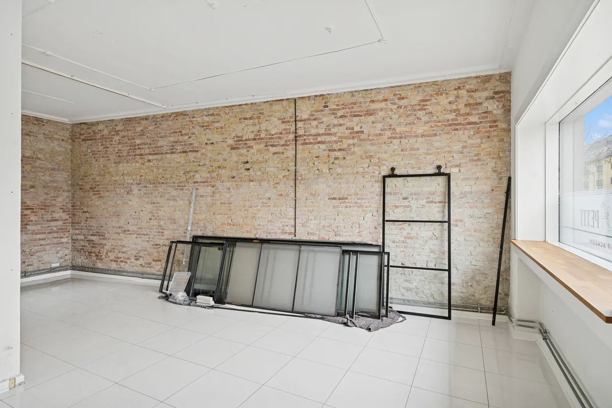 Tomme lokaler med hvide fliser og højloft. En slank stige og flere glasruder er placeret. Muren er lavet af mursten og der er et vindue med en udsigt til en bygning.