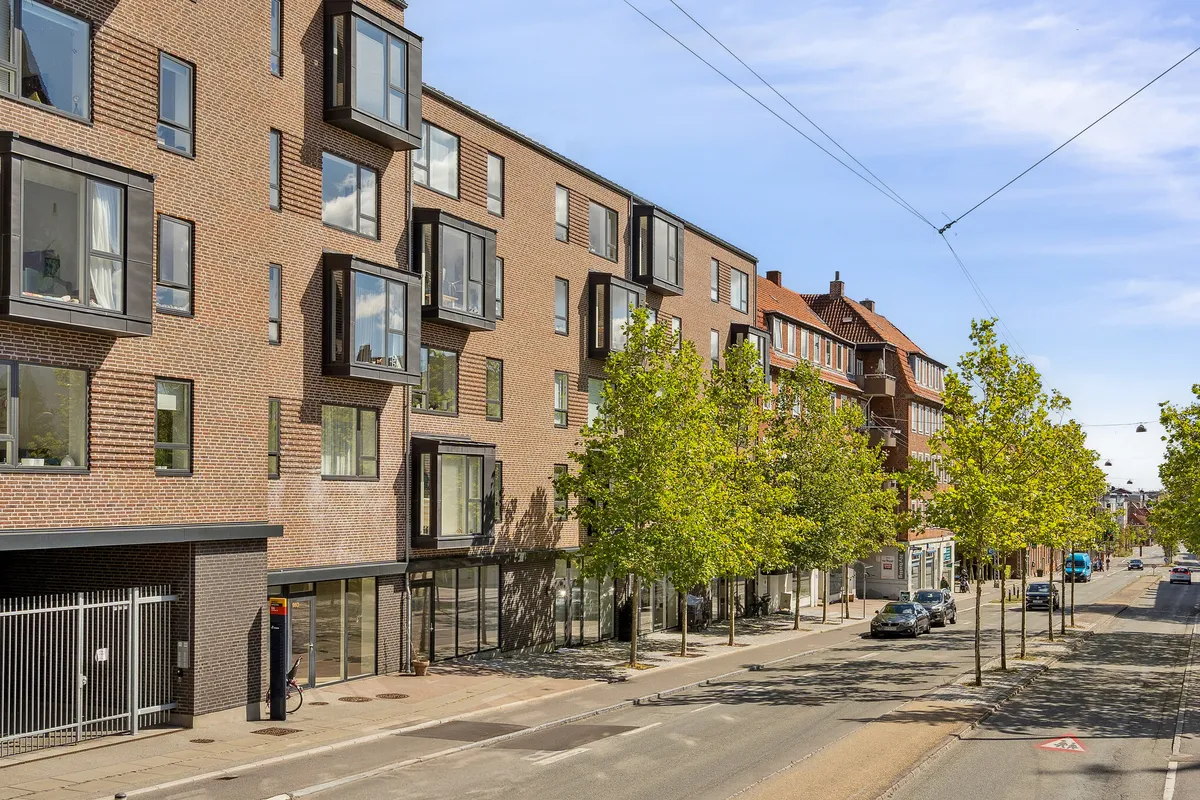 Moderne lejlighedsbygning med store glasvinduer og balkoner på Søborg Hovedgade. Bygningen er placeret ved siden af grønne træer og en travl gågade. På gågaden er der biler og busser.