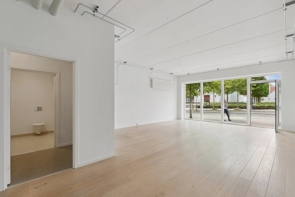 Et rum med hvide vægge og lyse trægulve. Glasser og en åben dør til et mindre rum med toilet. Udenfor ser man en gågade med grønne planter og buske