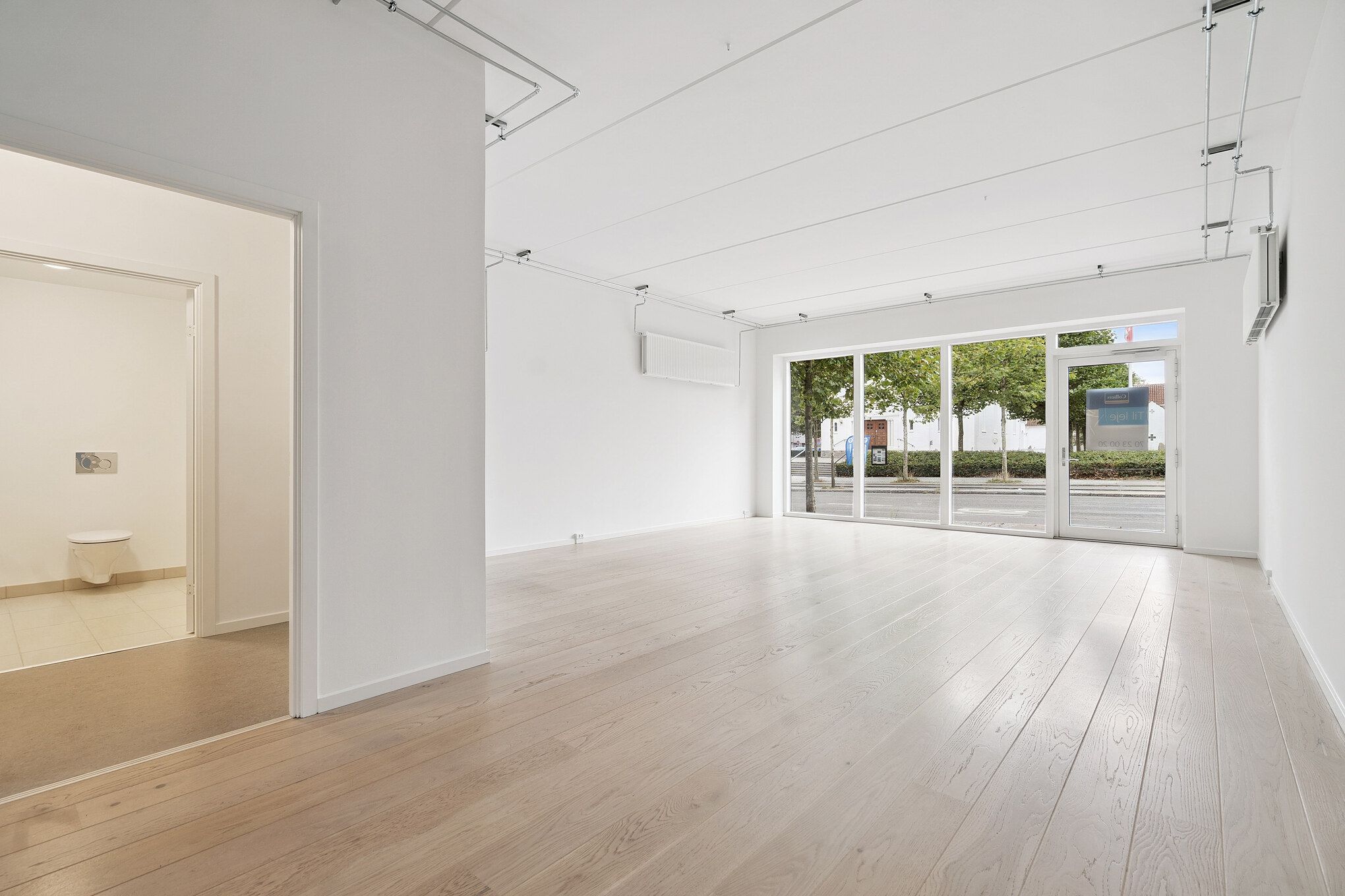 Et tomt lokale med lyse trægulve og glasdøre mod gaden. Der er hvide vægge og loft samt en toiletdør til venstre. Billedet viser en tydelig udsigt ud til en parkeret bil.