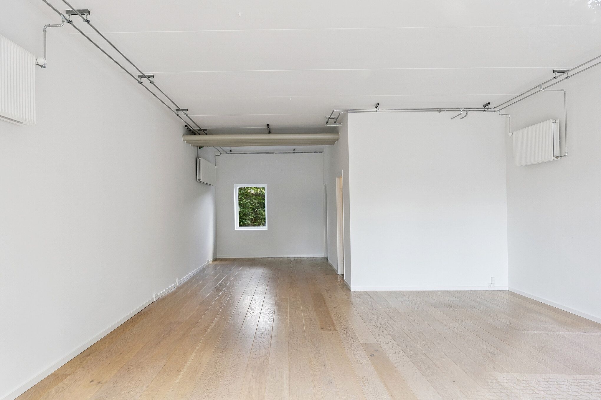 En stor, tom, hvidrum med trægulv og hvide vægge, der er ledsaget af hængende ventilationsanlæg. Rummet har et vindue med udsigt til grønne træer.