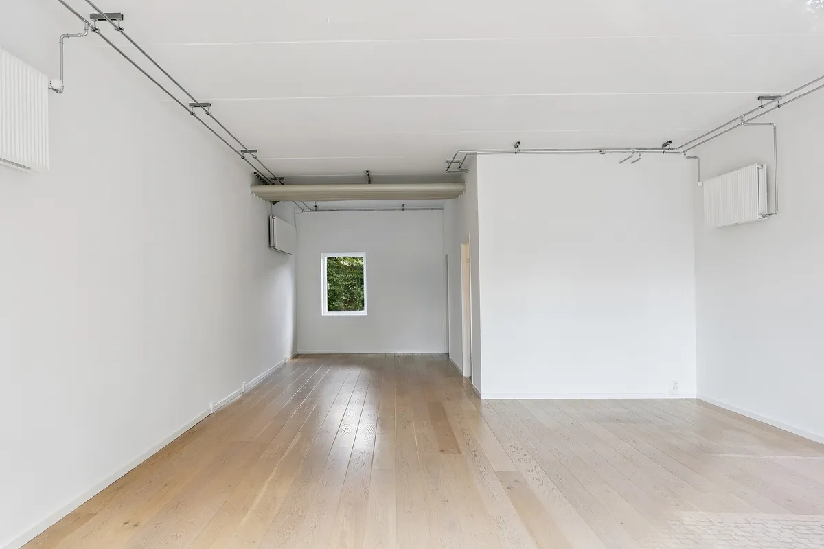 En stor, tom, hvidrum med trægulv og hvide vægge, der er ledsaget af hængende ventilationsanlæg. Rummet har et vindue med udsigt til grønne træer.