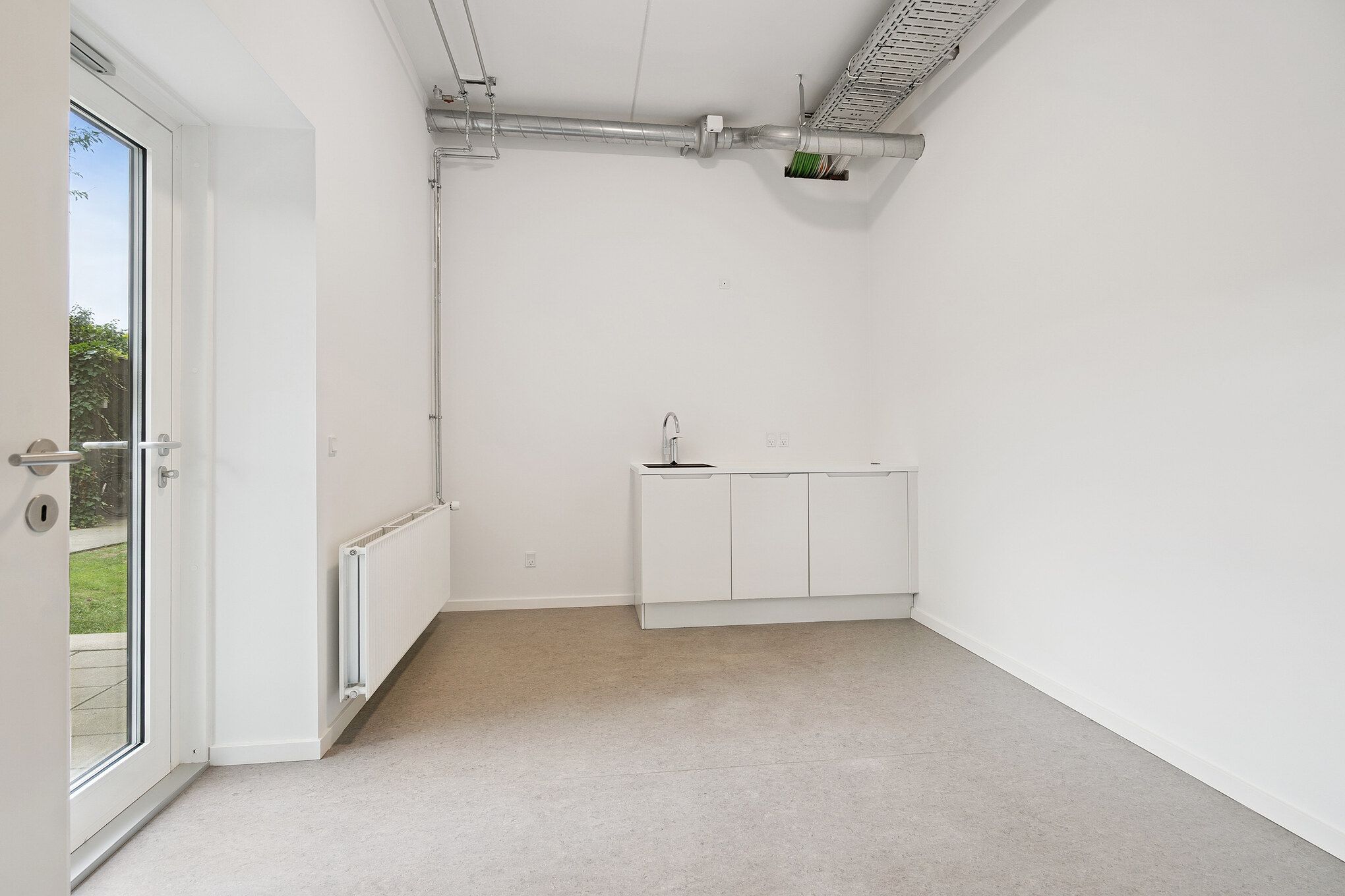 Et tomt lokale med hvide vægge og gulv, hvor der er et vindue, døre og en hvid radiator, samt en ren, hvid og funktionel køkkenskab.