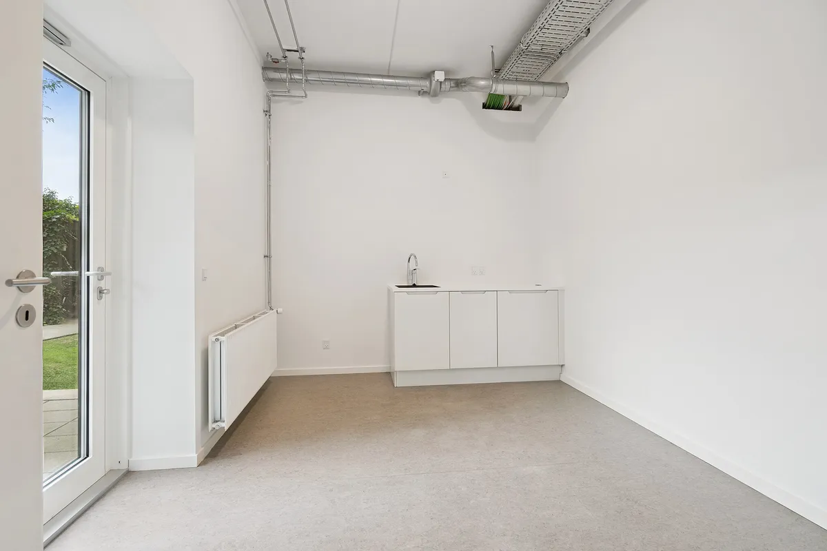 Et tomt lokale med hvide vægge og gulv, hvor der er et vindue, døre og en hvid radiator, samt en ren, hvid og funktionel køkkenskab.