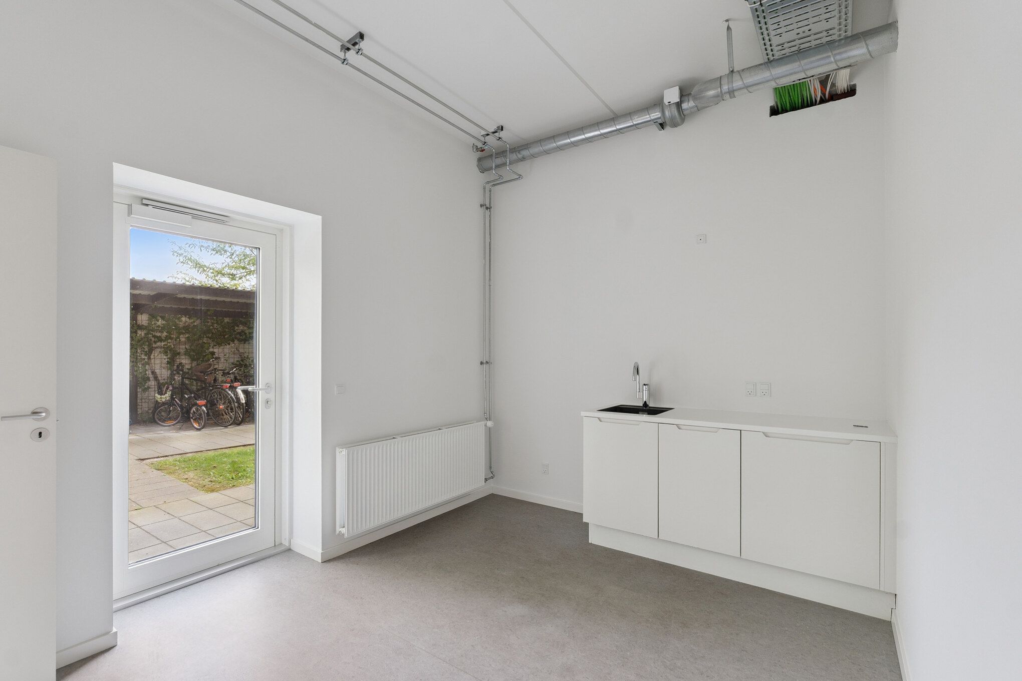 Rummelig kontorplads med hvid væg og hvidt gulv samt køkken med sink og håndklæder. Der er et vindue med glasdør og udsigt til græsplæne med cykler.