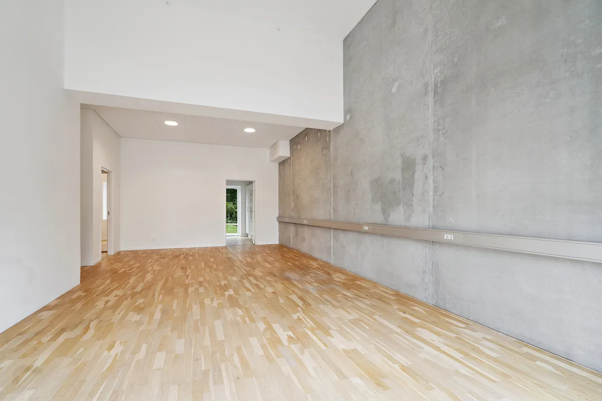 Et stort, tomt rum med højt hvide loft og vægge samt lysegrå betonvæg og lysegrå trægulv. To døre åbner sig mod andre rum og udendørs
