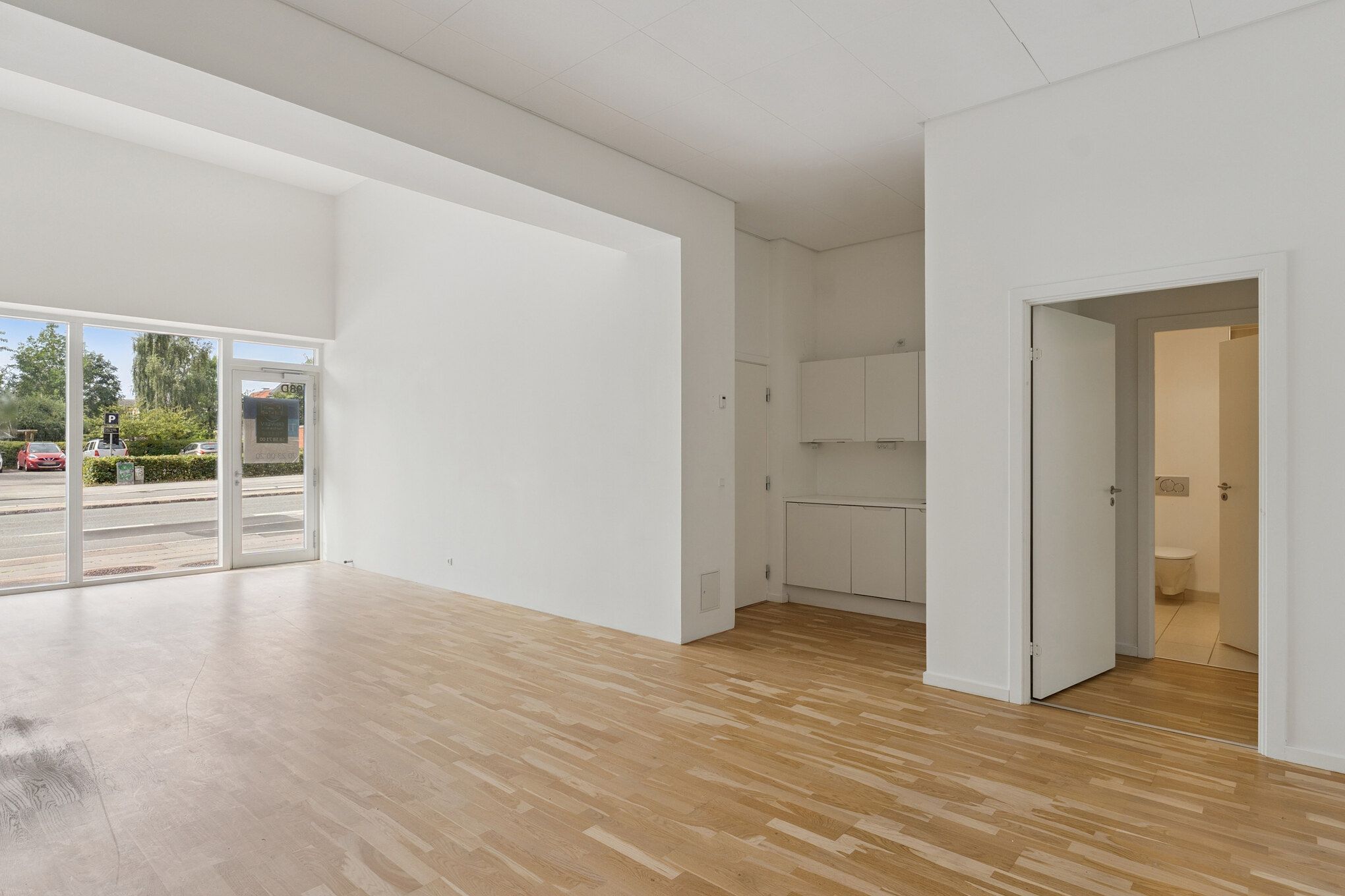 Funktionelt erhvervslejemål med store vinduer og højt loft, lys og moderne design, hvidt køkken med lyse skabsdøre, indgangsdør til toiletrum og parkeringsplads udenfor.