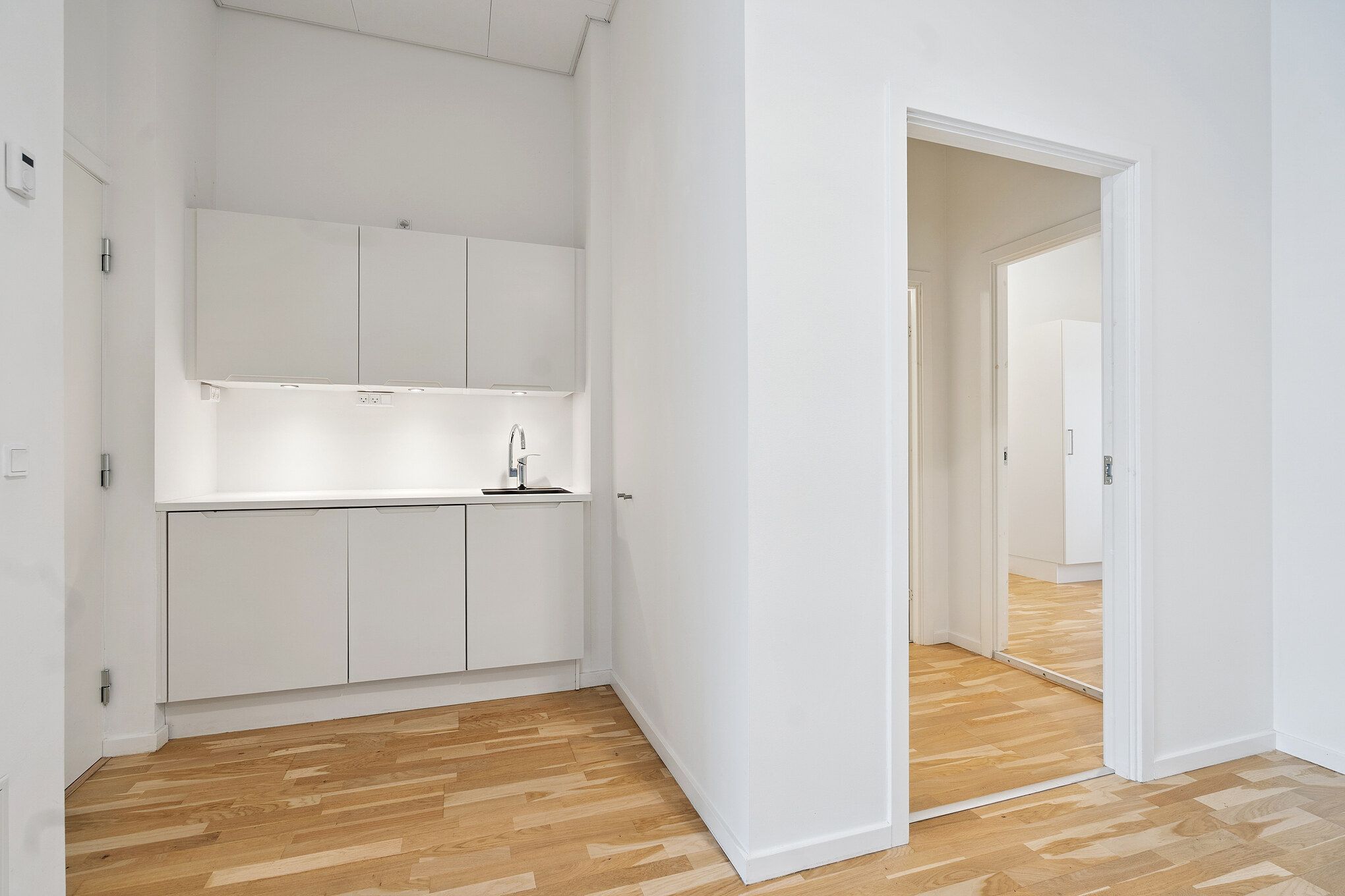 En moderne og minimalistisk køkken med hvide vægge og trægulv, der skaber en ren og funktionel atmosfære. Der er lysekroner og hængeskabe, og et spejl reflekterer det tomme rum