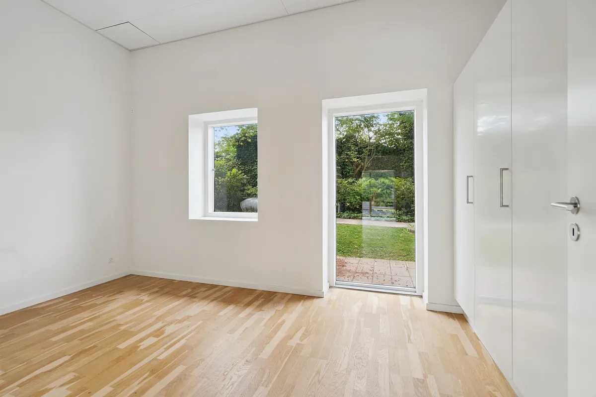 Et tomt rum med lyse hvide vægge og gulvtæppe, to vinduer og en glasdør med håndtag og lås til en have med græsplæne og grønne planter