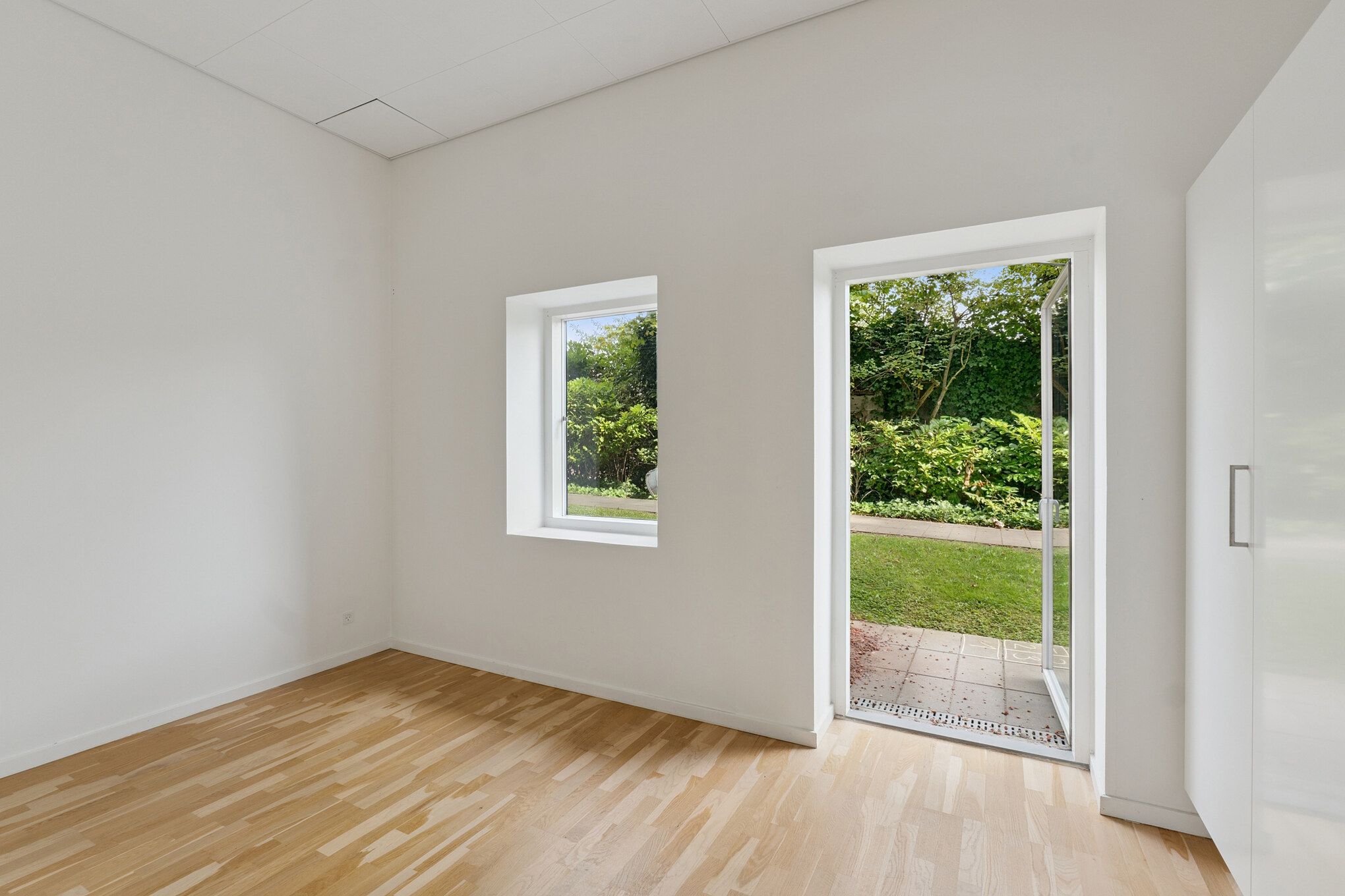 Et rum med lyse vægge og trægulv, hvor en dør står åben mod en have med grønne planter og græs. To vinduer med hvide rammer giver udsigt til træerne udenfor