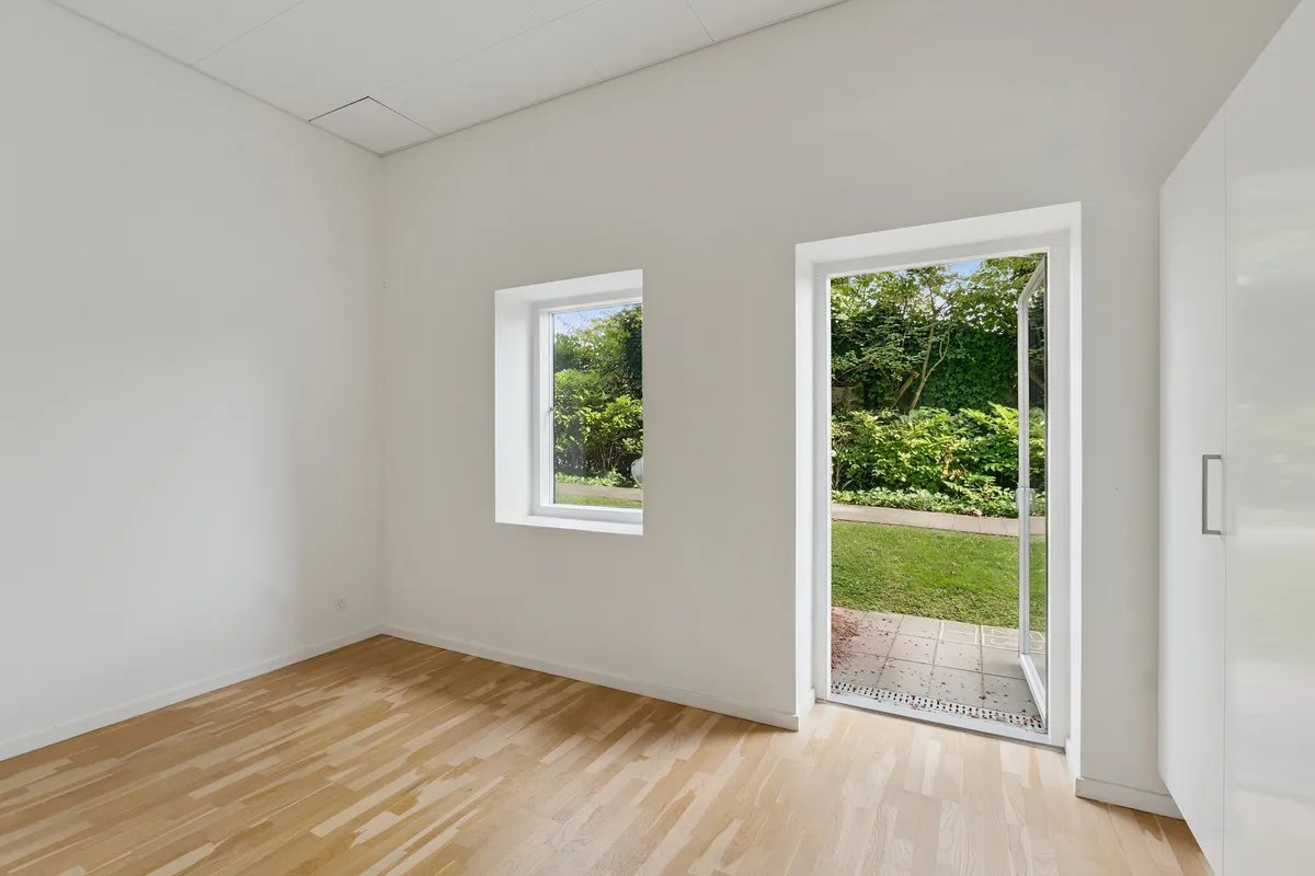 Et rum med lyse vægge og trægulv, hvor en dør står åben mod en have med grønne planter og græs. To vinduer med hvide rammer giver udsigt til træerne udenfor